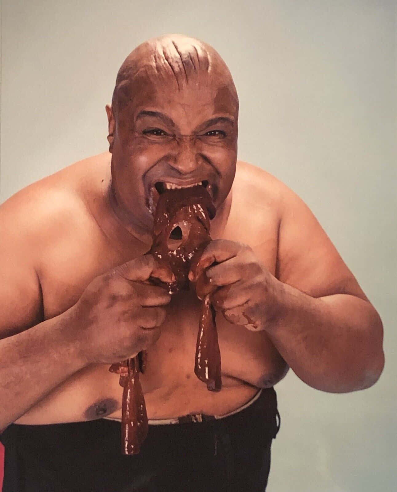 Abdullah The Butcher 桌布 壁紙 — 一個光頭男人帶著喜悅的表情，雙手拿著一個大的巧克力覆蓋的糕點咬了一口。