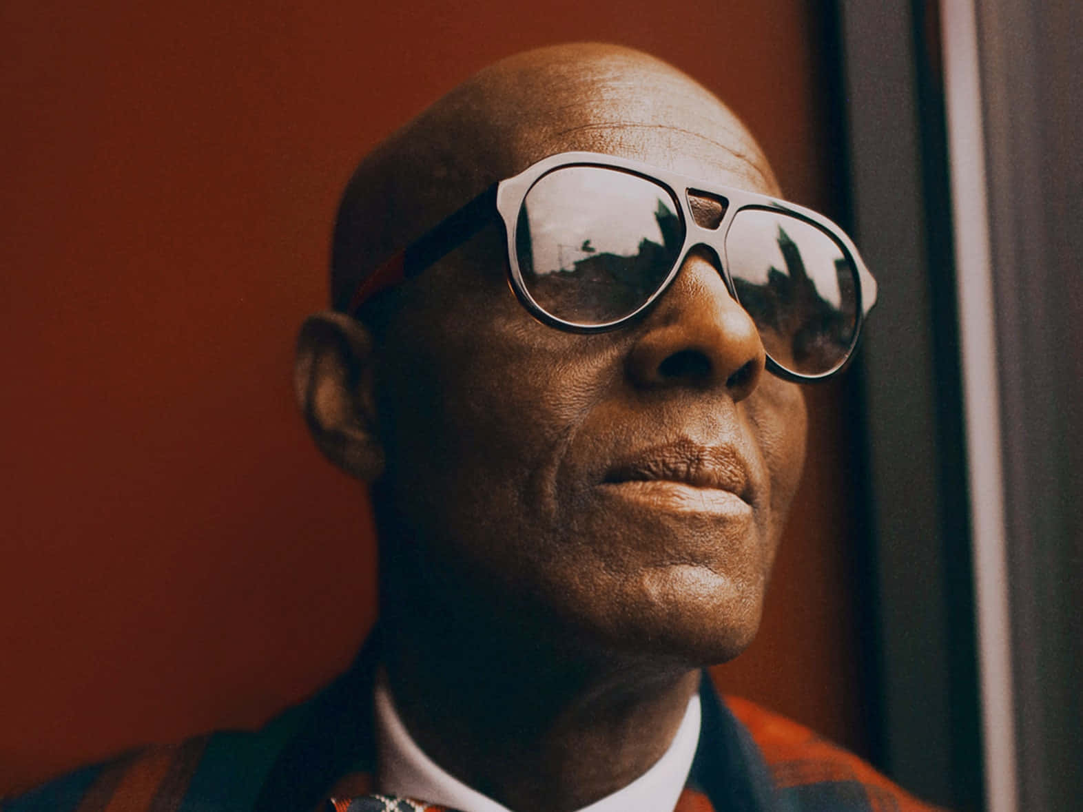 Sunglasses 桌布 壁紙 — 一個光頭男人戴着超大太陽眼鏡，對著溫暖的紅牆，若有所思地望向窗外。