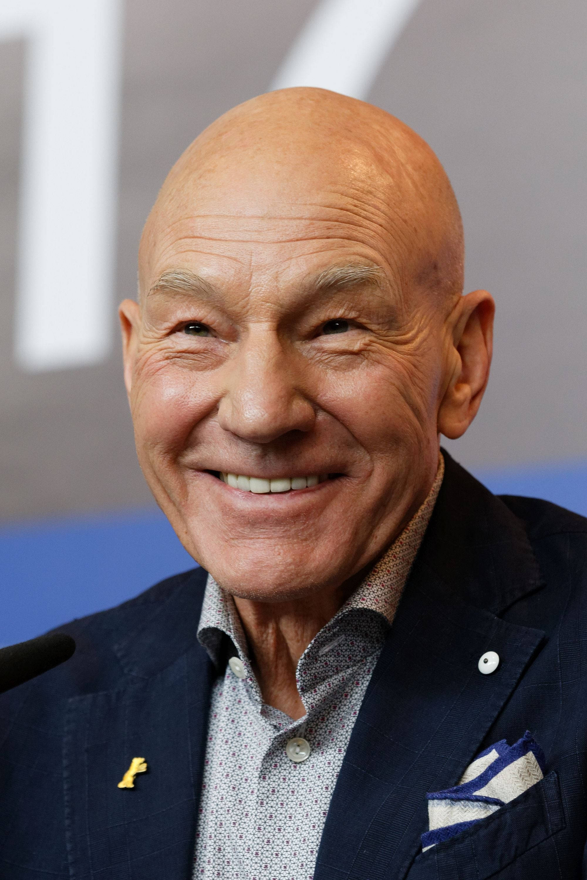 Celebrity 桌布 壁紙 — 一個光頭男人的特寫肖像，他有深深的皺紋，穿著深色西裝和圖案襯衫，溫暖地微笑。