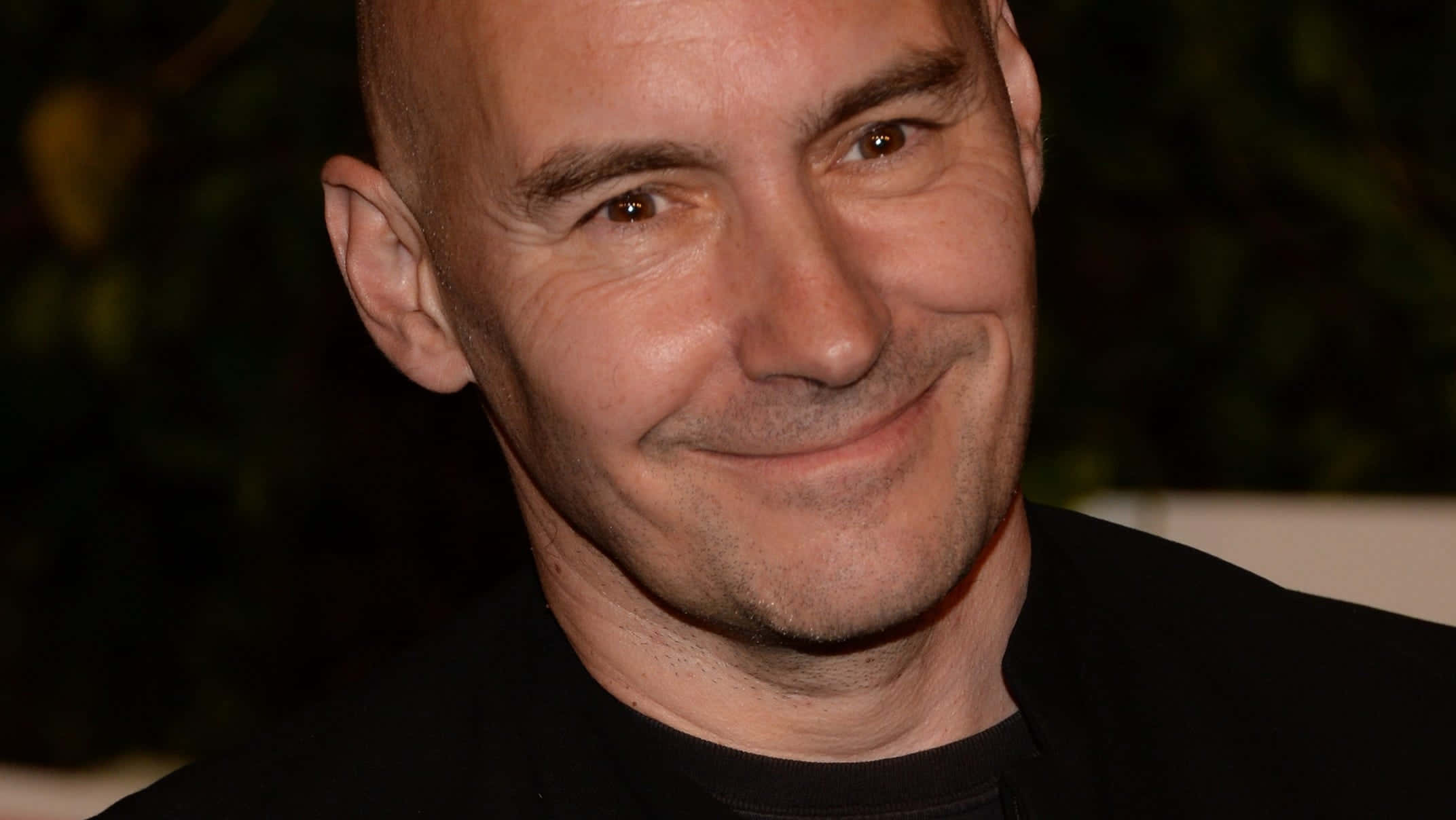 Grant Morrison 桌布 壁紙 — 一個光頭男人的近距離肖像，帶著溫暖的笑容，穿著黑色襯衫，背景是暗色模糊的。