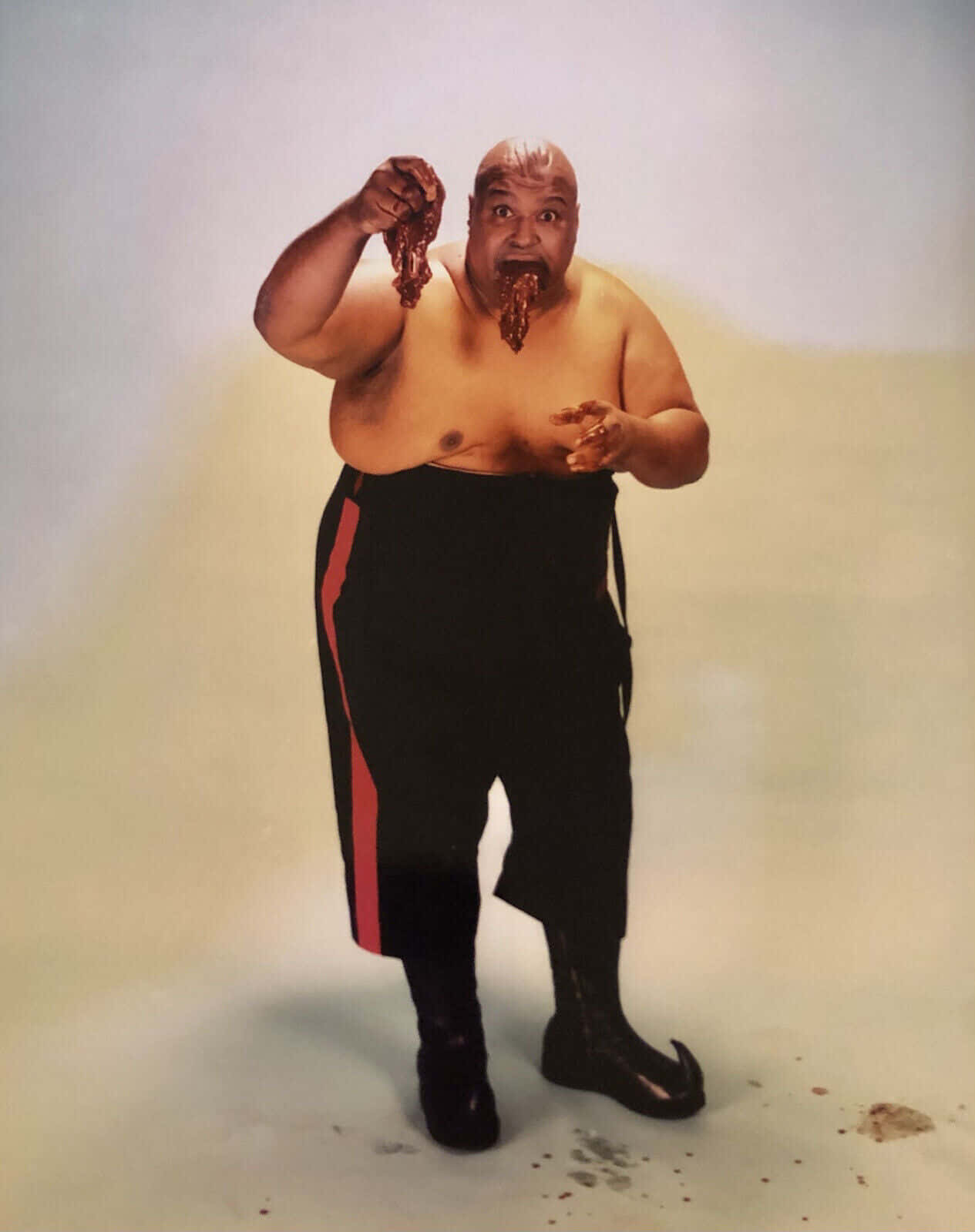 WWE 桌布 壁紙 — 一個光頭男人穿著帶有紅色條紋的黑色褲子，站在素色背景前，拿著並吃著一大塊肉，醬汁從他的嘴裡滴下來。