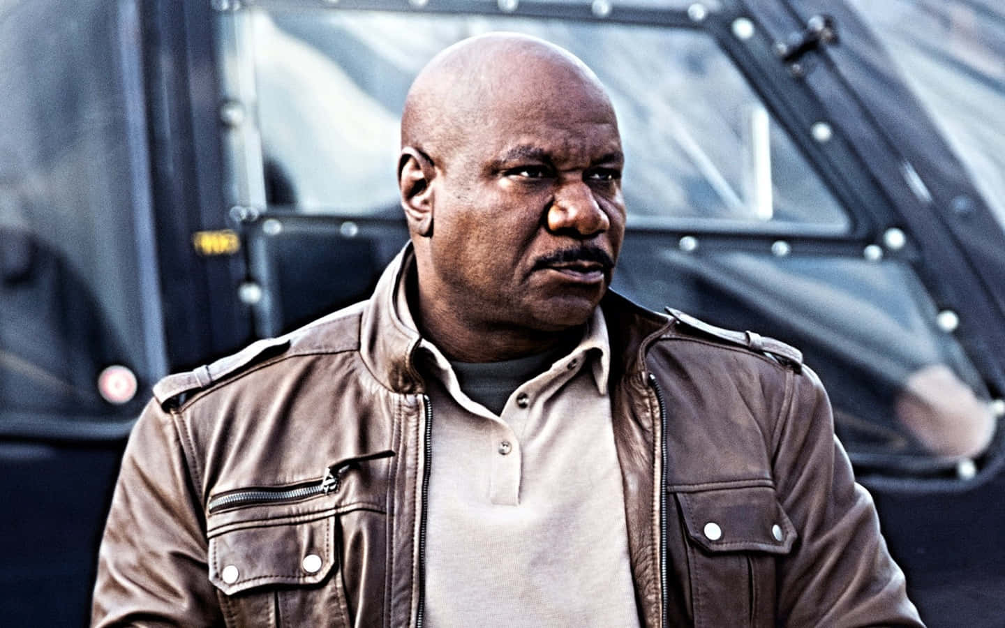 Ving Rhames 桌布 壁紙 — 一個光頭男人穿著棕色皮夾克，神情嚴肅地站在一輛黑色裝甲車前。
