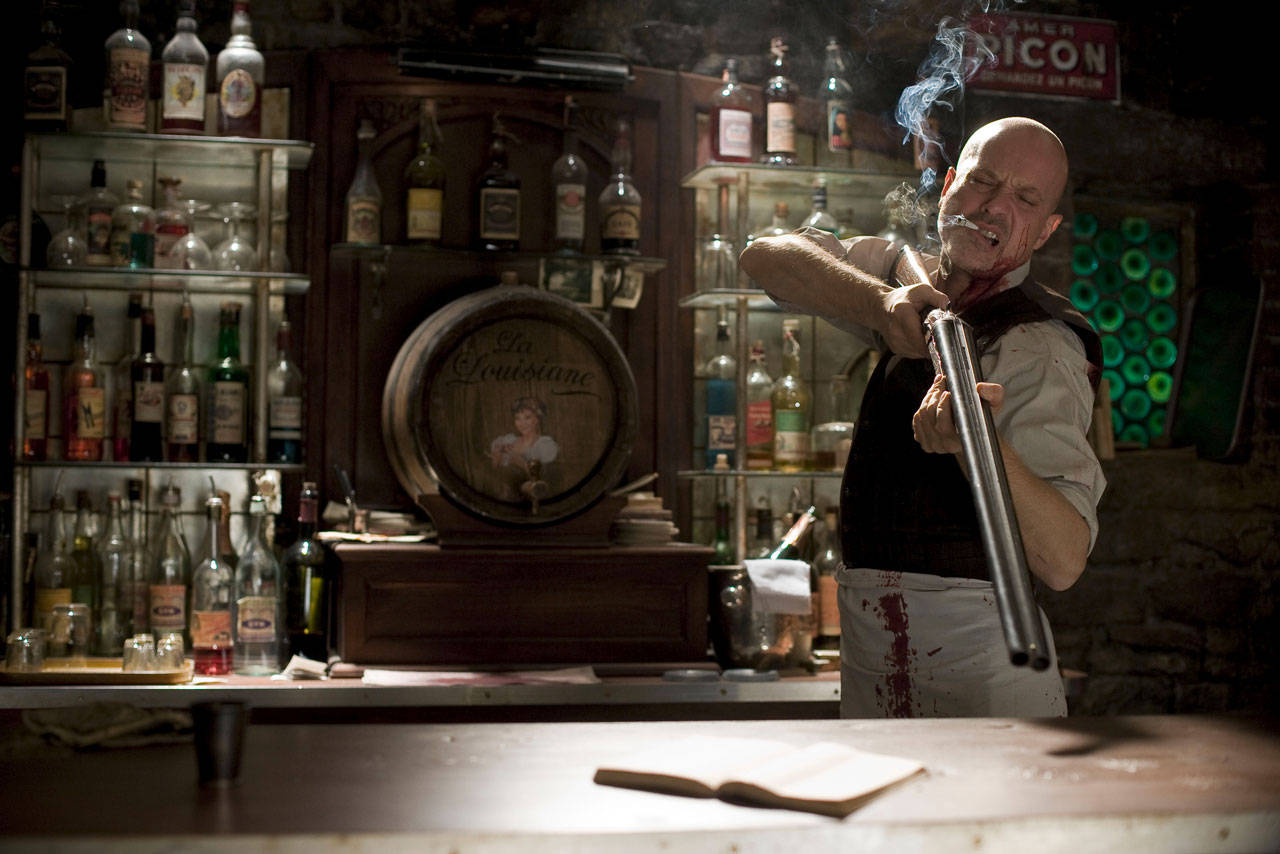 Inglourious Basterds 桌布 壁紙 — 一個光頭男人穿著沾滿血跡的圍裙，在燈光昏暗的酒吧裡抽著雪茄，手持霰彈槍，身後有酒瓶和一個標有「La Louisiane」的木桶。