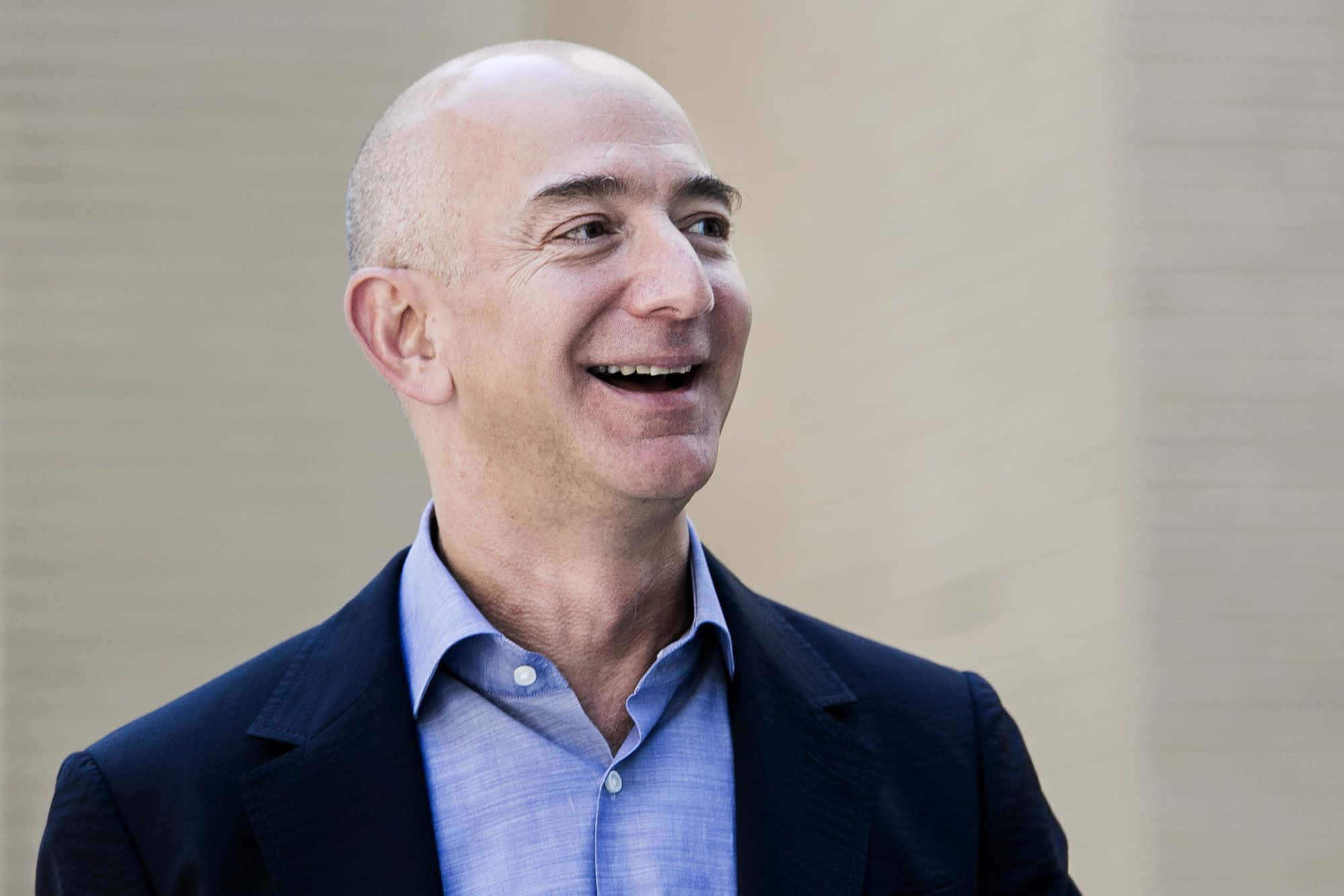 Jeff Bezos 桌布 壁紙 — 一個光頭男人穿著深色西裝和藍色襯衫，對著純米色牆壁開懷大笑。