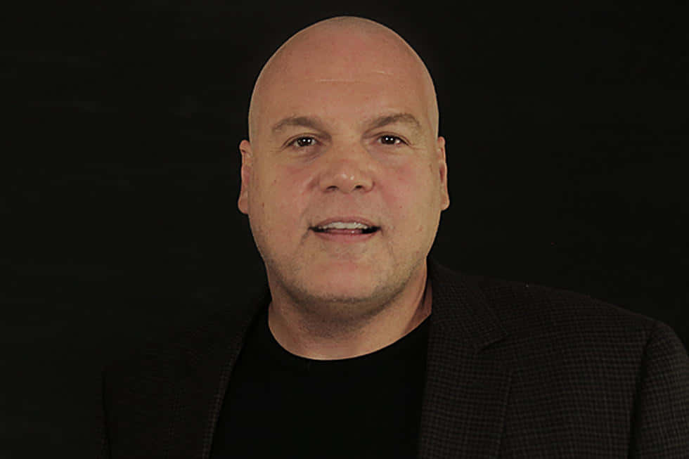 Vincent D'onofrio 桌布 壁紙 — 一個光頭男人穿著深色西裝和黑色襯衫，在純黑色背景上溫柔地微笑。