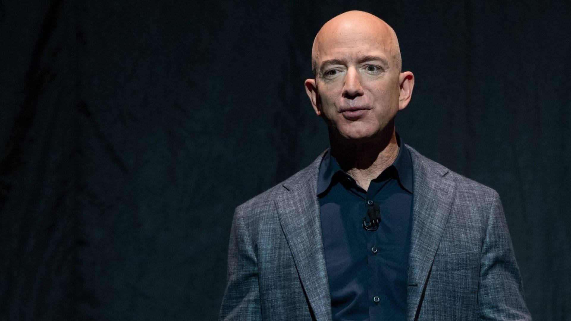 Bezos 桌布 壁紙 — 一個光頭男人穿著灰色格紋西裝和深色襯衫，在舞台燈光下站在深色紋理背景前。