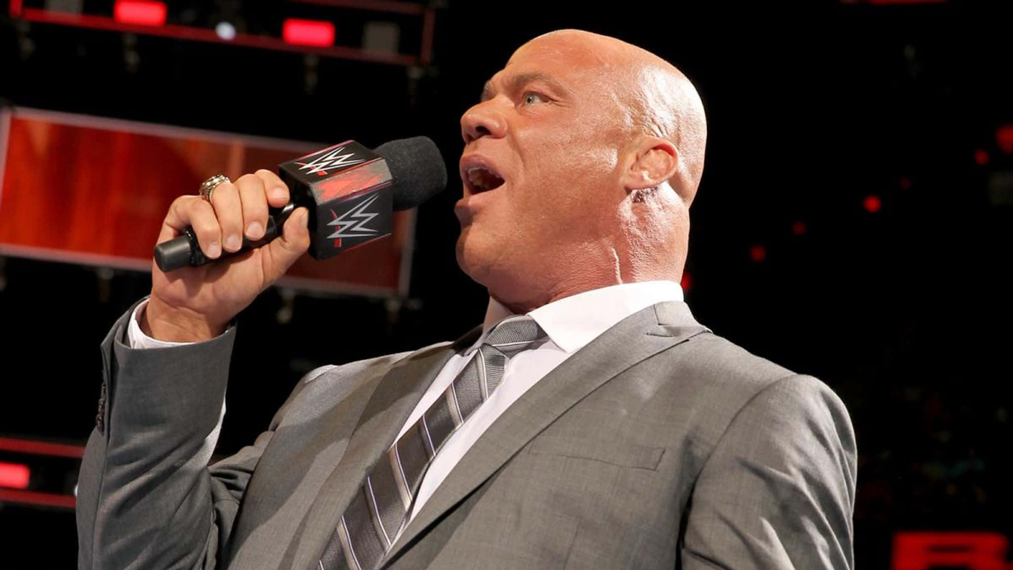 WWE 桌布 壁紙 — 一個光頭男人穿著灰色西裝，在明亮的舞台燈光下熱情地對著WWE麥克風說話。