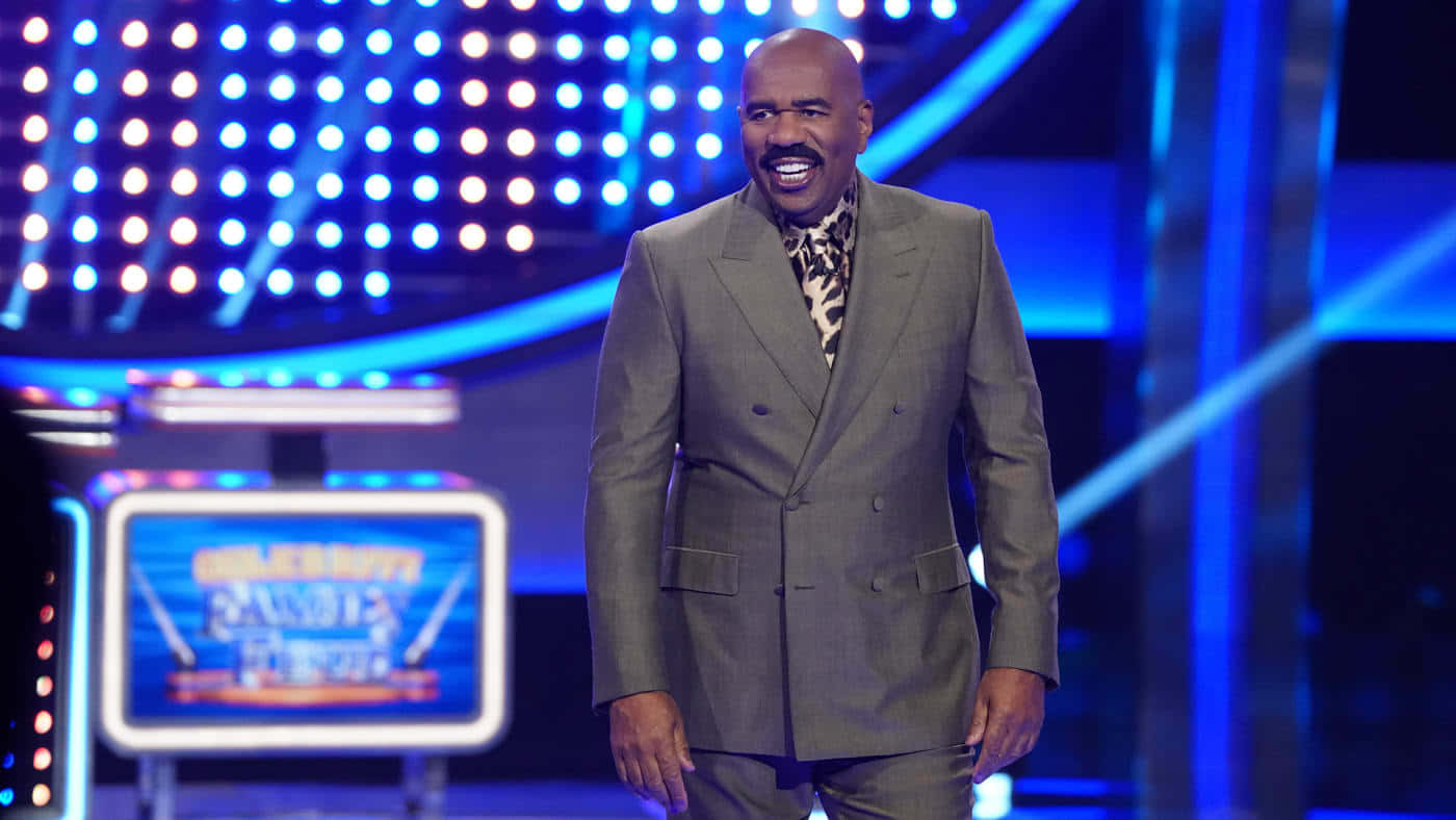 steve harvey 桌布 壁紙 — 一個光頭男人穿著灰色西裝和圖案領帶，在一個燈光明亮、有藍色燈光和「Celebrity Name That Tune」螢幕的遊戲節目舞台上開懷大笑。