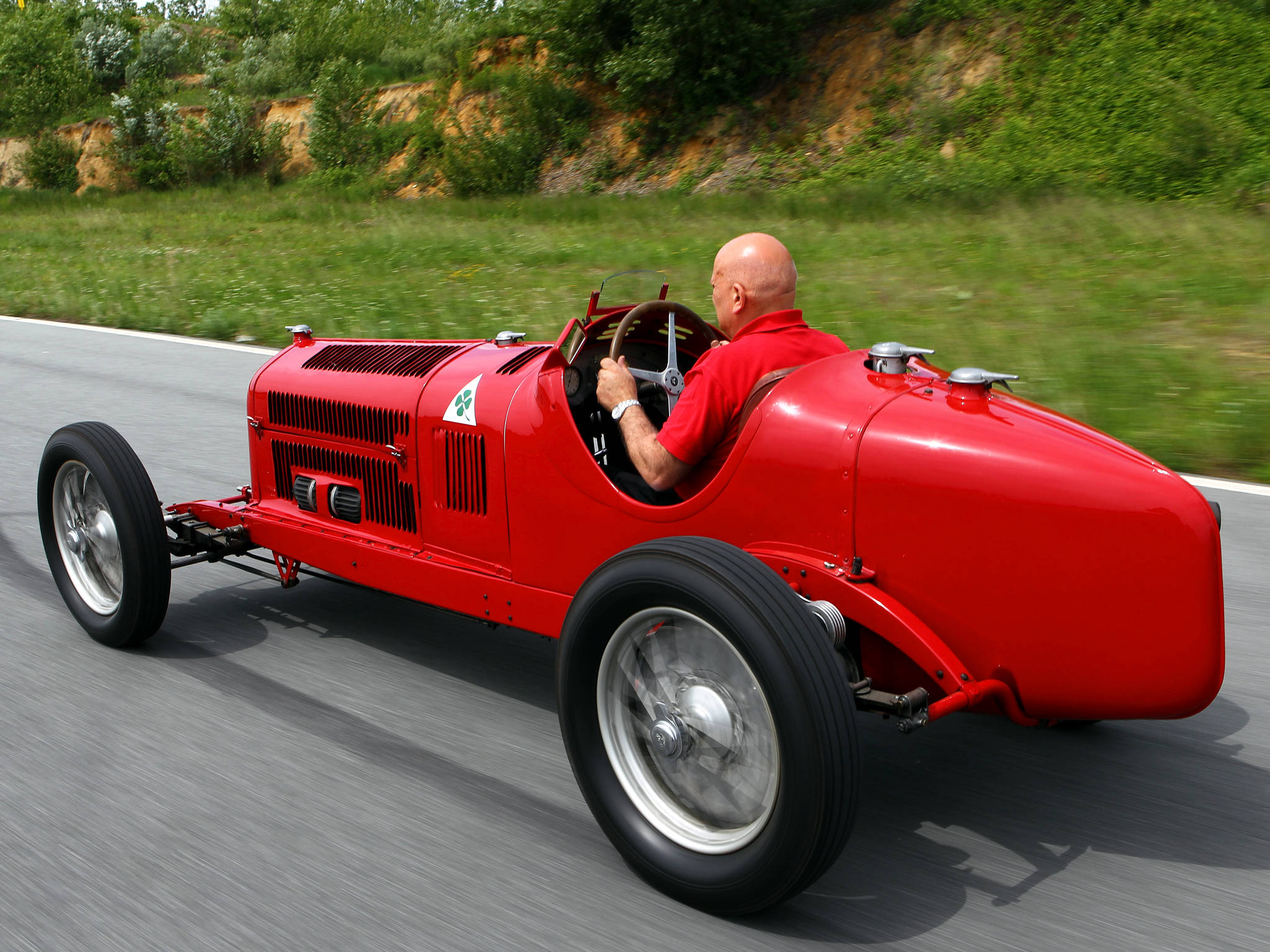 alfa romeo iPhone 桌布 壁紙 — 一個光頭男人穿著紅色襯衫，駕駛一輛復古紅色賽車沿著蜿蜒的道路穿過鬱鬱蔥蔥的綠色山丘。