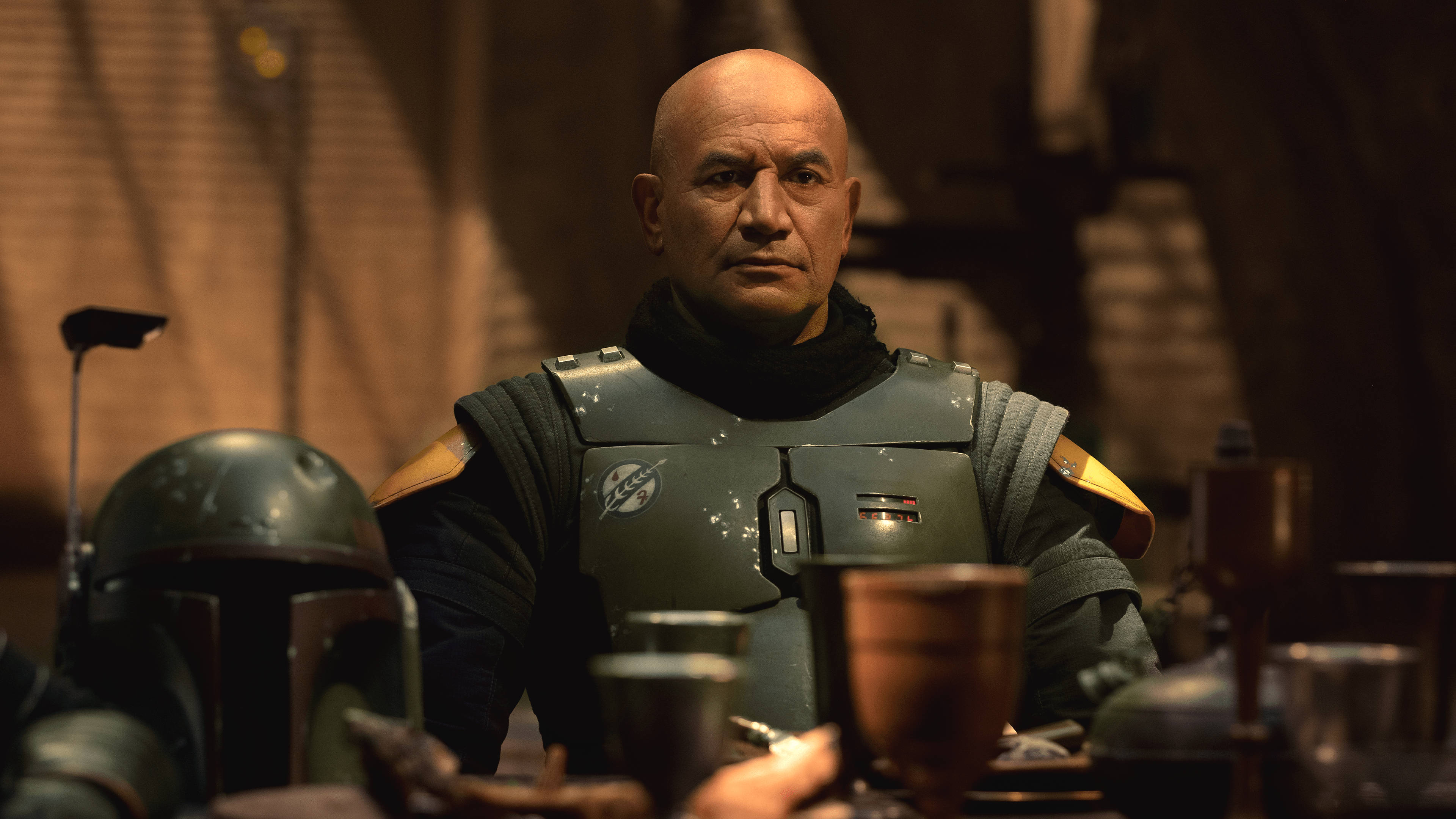 Boba 桌布 壁紙 — 一個光頭男人穿著風化盔甲，坐在昏暗的桌子旁，頭盔放在旁邊，身處一個粗糙的酒館。