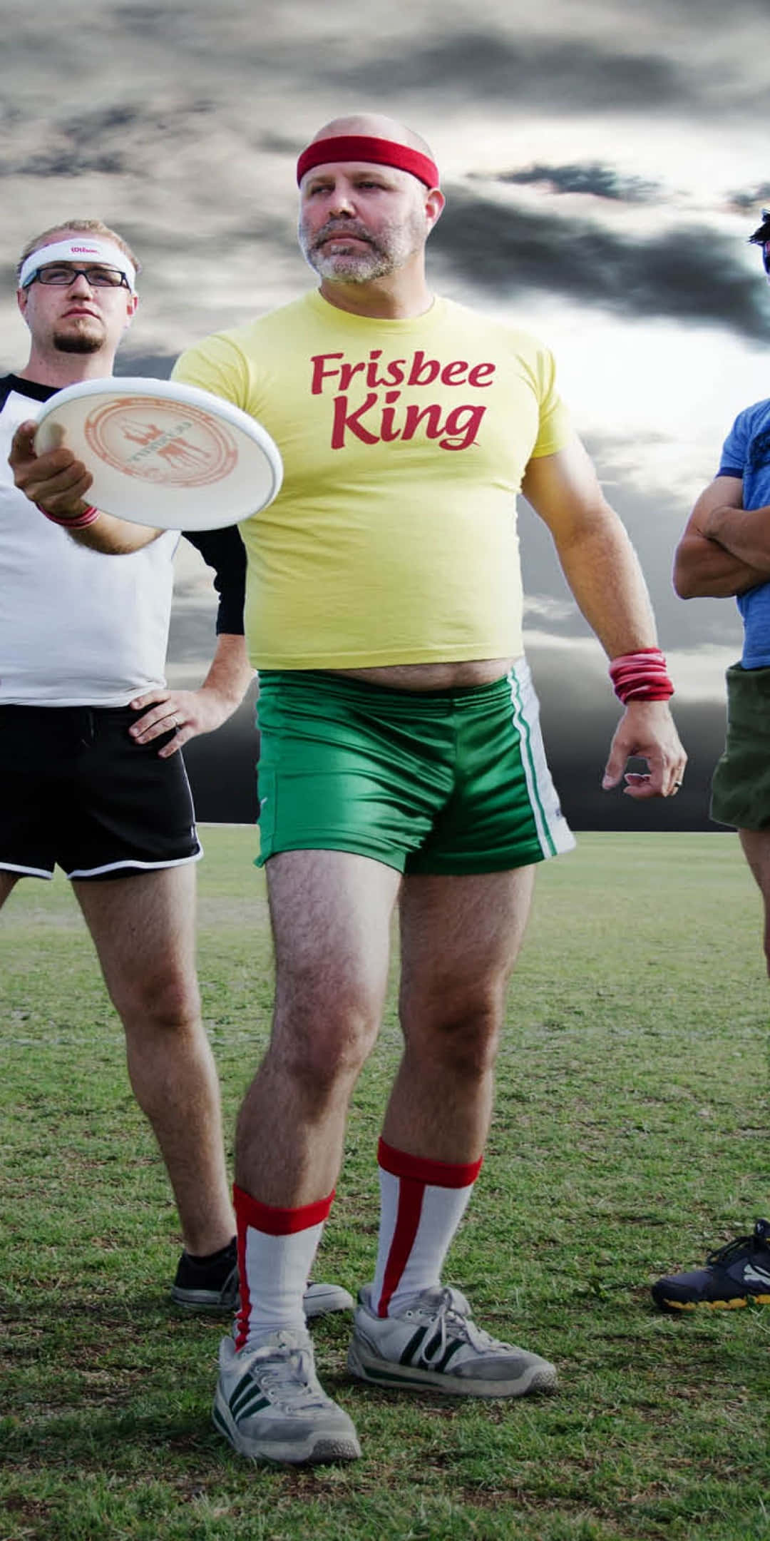 Pixel 3 Frisbee 桌布 壁紙 — 一個光頭男人穿著黃色「Frisbee King」T恤和綠色短褲，自信地站在草地上，在陰天中拿著飛盤。