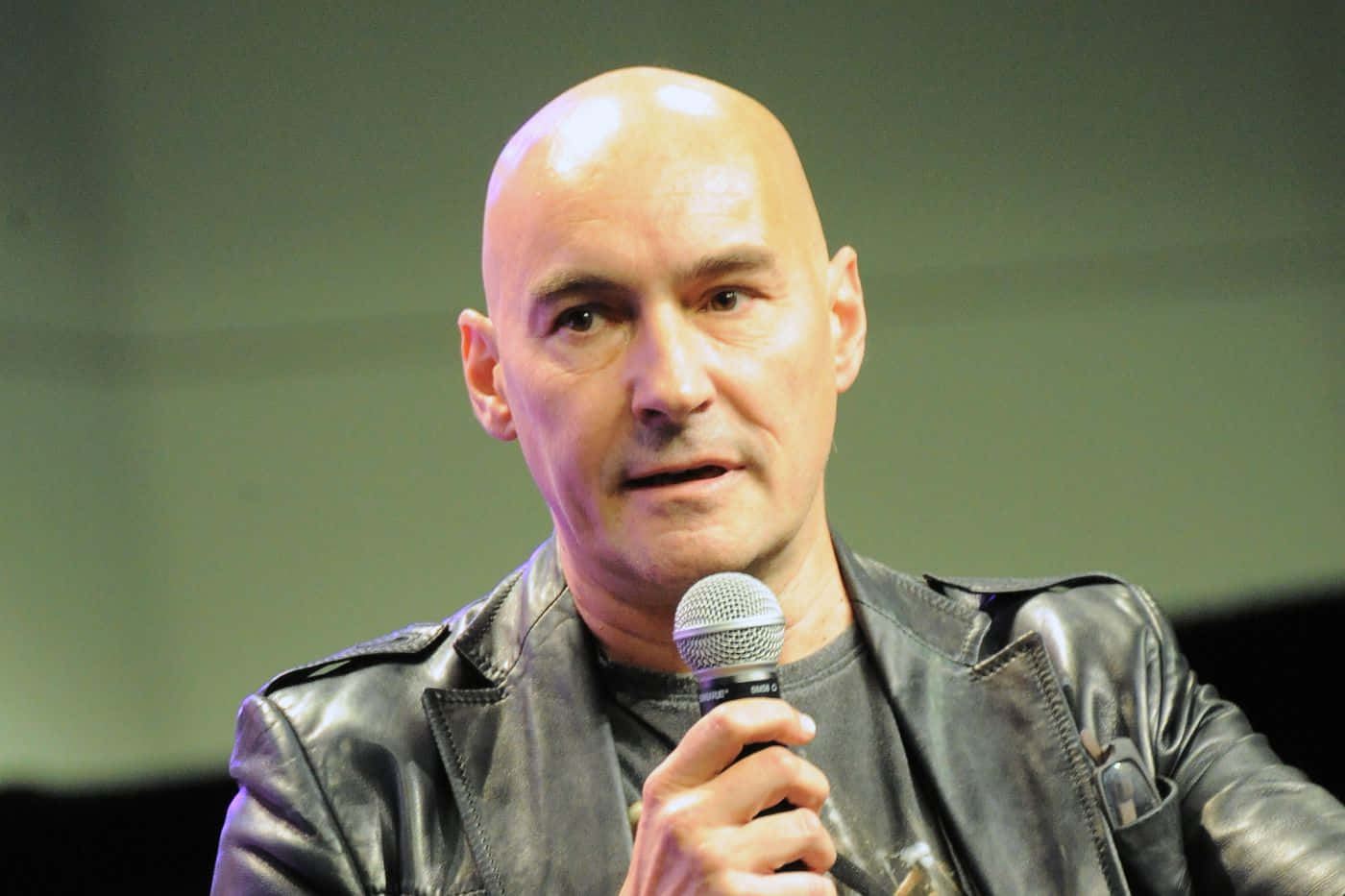 Grant Morrison 桌布 壁紙 — 一個光頭男人穿著黑色皮夾克，專注地對著麥克風說話。