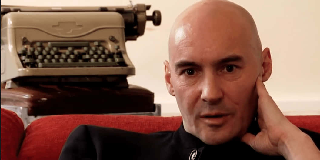 Grant Morrison 桌布 壁紙 — 一個光頭男人穿著黑色襯衫，手托下巴坐在紅色沙發上，身後有一台復古米色打字機。