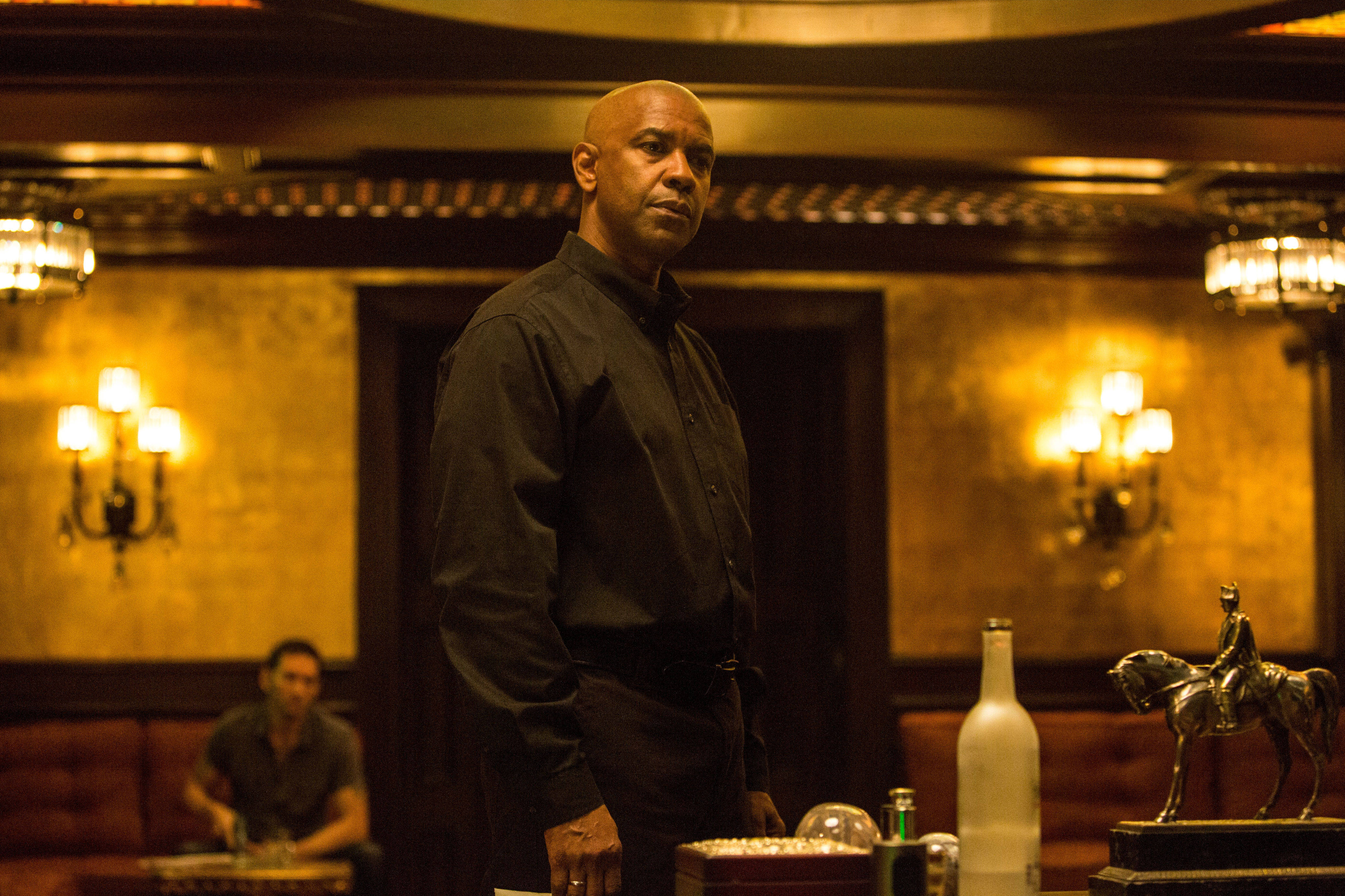 Denzel Washington 桌布 壁紙 — 一個光頭男人穿著黑色襯衫，莊嚴地站在一個溫暖燈光、華麗的酒吧裡，有金色牆壁和復古壁燈。