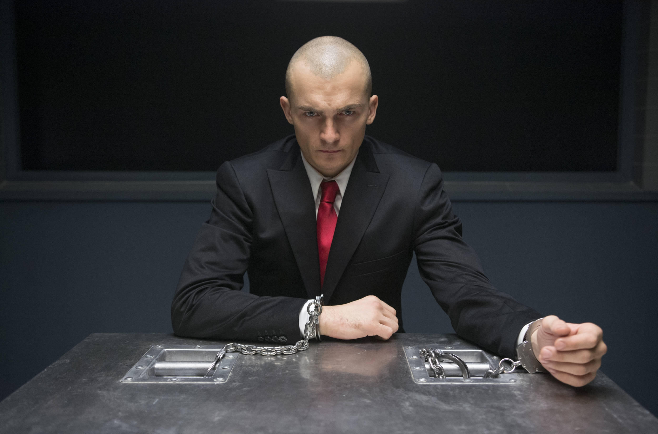 Hitman Agent 47 桌布 壁紙 — 一個光頭男人穿著黑色西裝和紅色領帶，被銬在一個簡陋審訊室的金屬桌上。