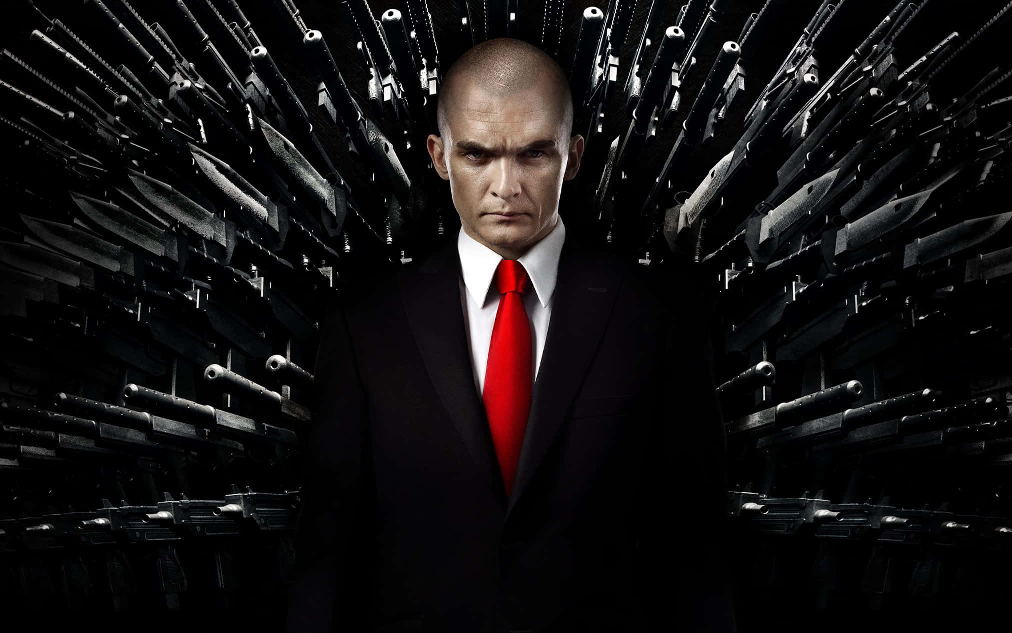 Hitman Desktop 桌布 壁紙 — 一個光頭男人穿著黑色西裝和紅色領帶，表情嚴肅地站在中央，被放射狀排列的武器包圍在深色背景前。