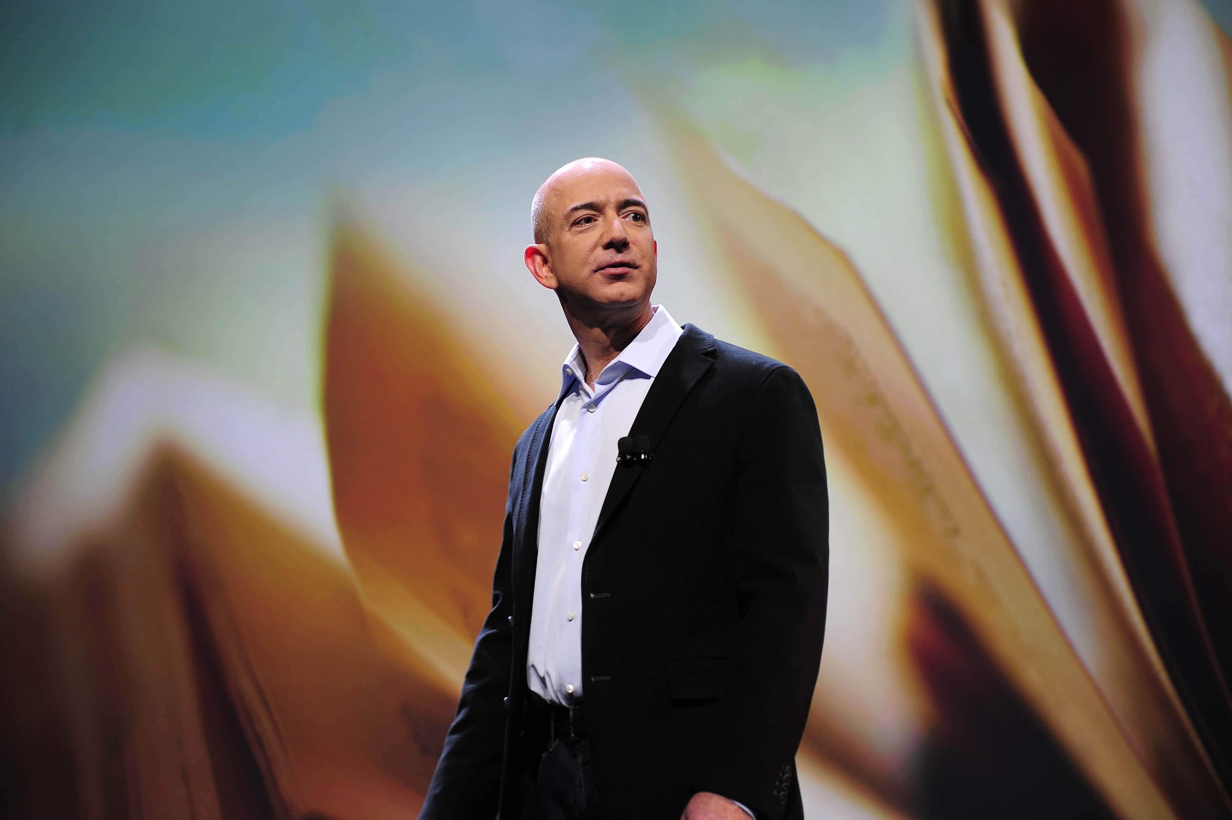 Jeff Bezos 桌布 壁紙 — 一個光頭男人穿著黑色西裝自信地站在舞台上，背景是模糊的抽象金色和藍綠色。
