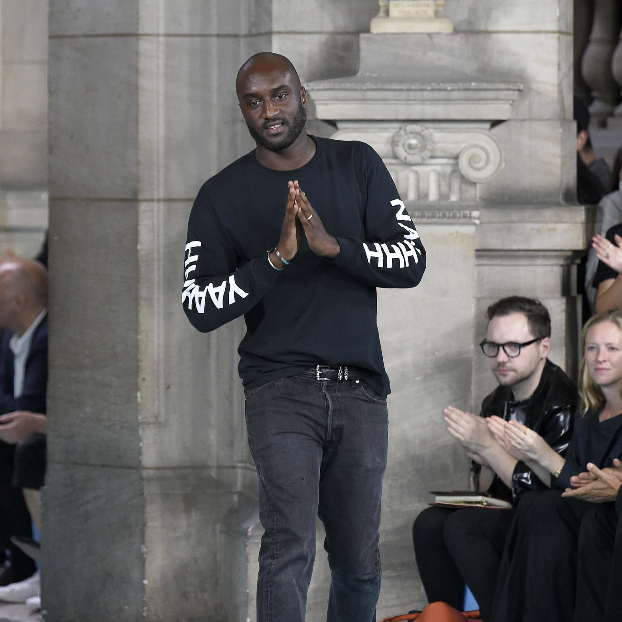 Off White 桌布 壁紙 — 一個光頭男人穿著黑色長袖衫，袖上有白色文字，站在石柱前拍手，周圍的人也在拍手。