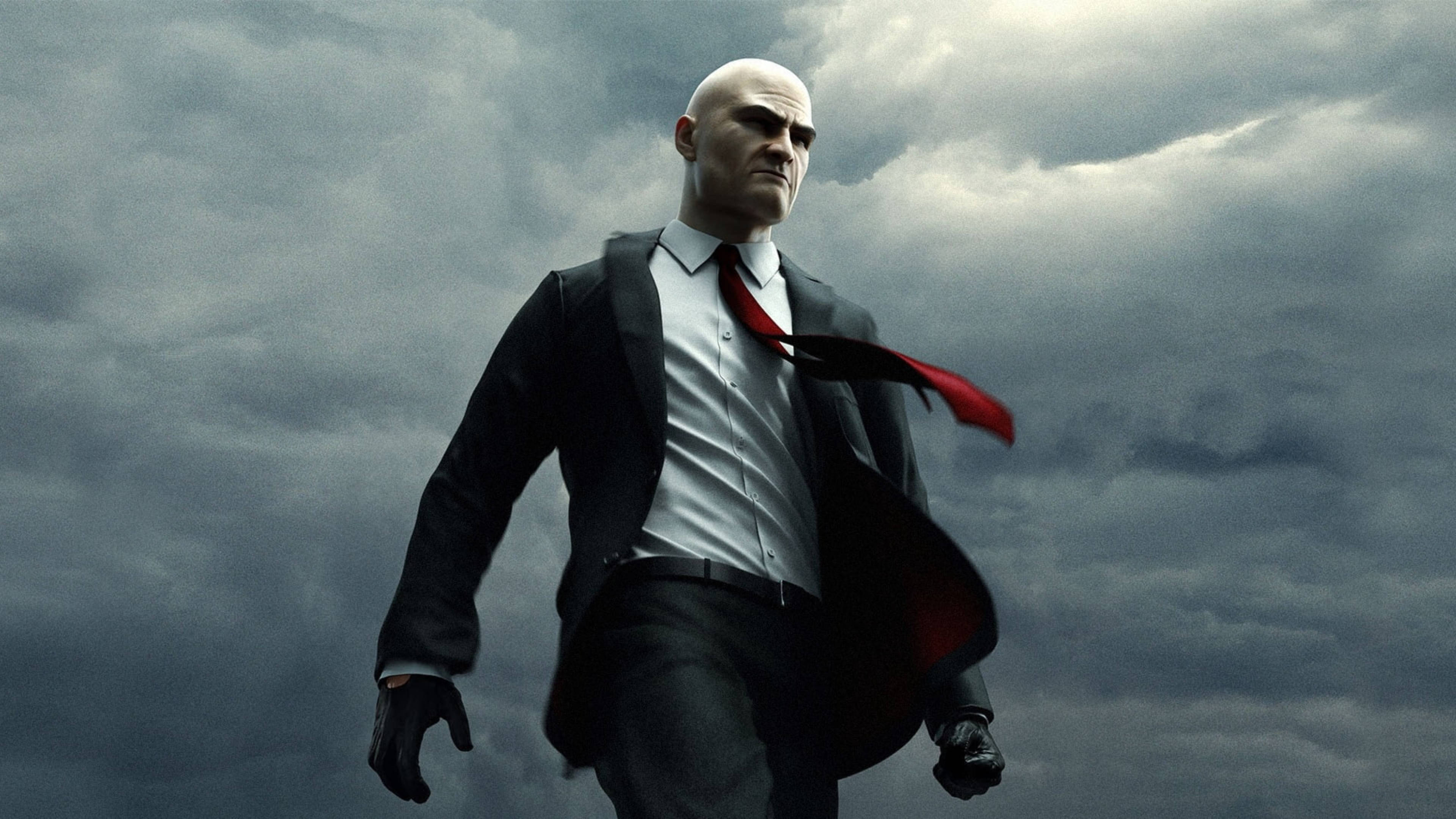 桌布 壁紙 — 一個光頭男人身穿黑色西裝和紅色領帶，在風暴般的灰色天空下大步向前。