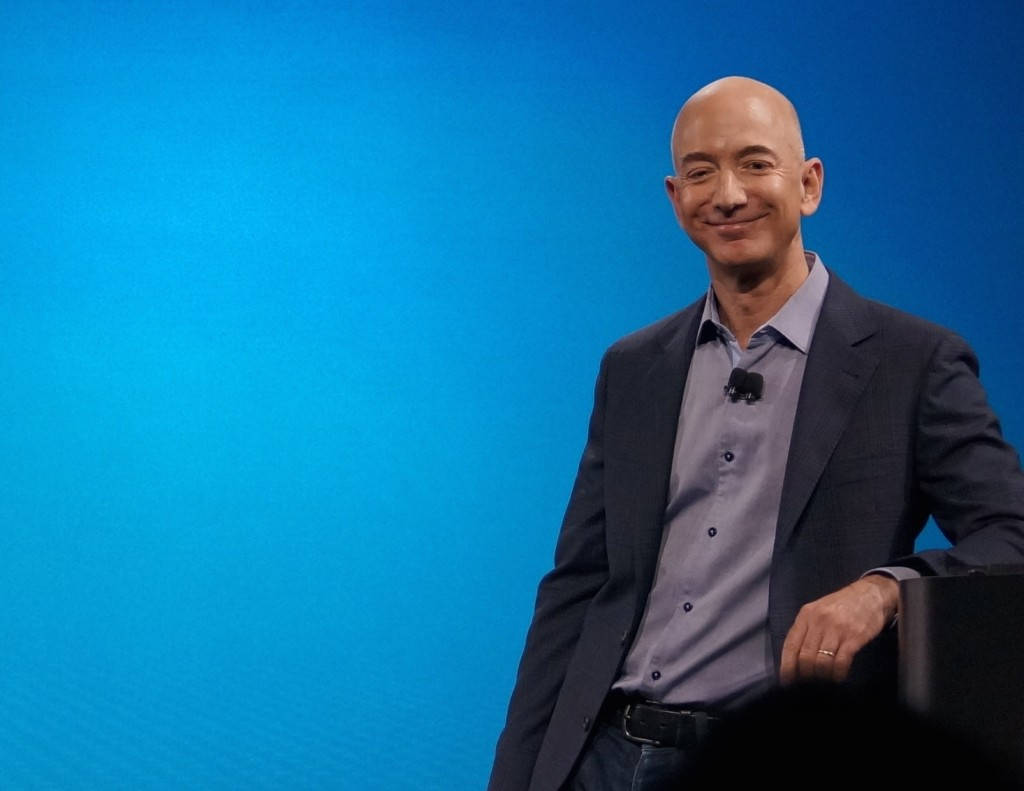 Bezos 桌布 壁紙 — 一個光頭男士穿著深色西裝和淺藍色襯衫，在純藍色背景前溫暖地微笑。