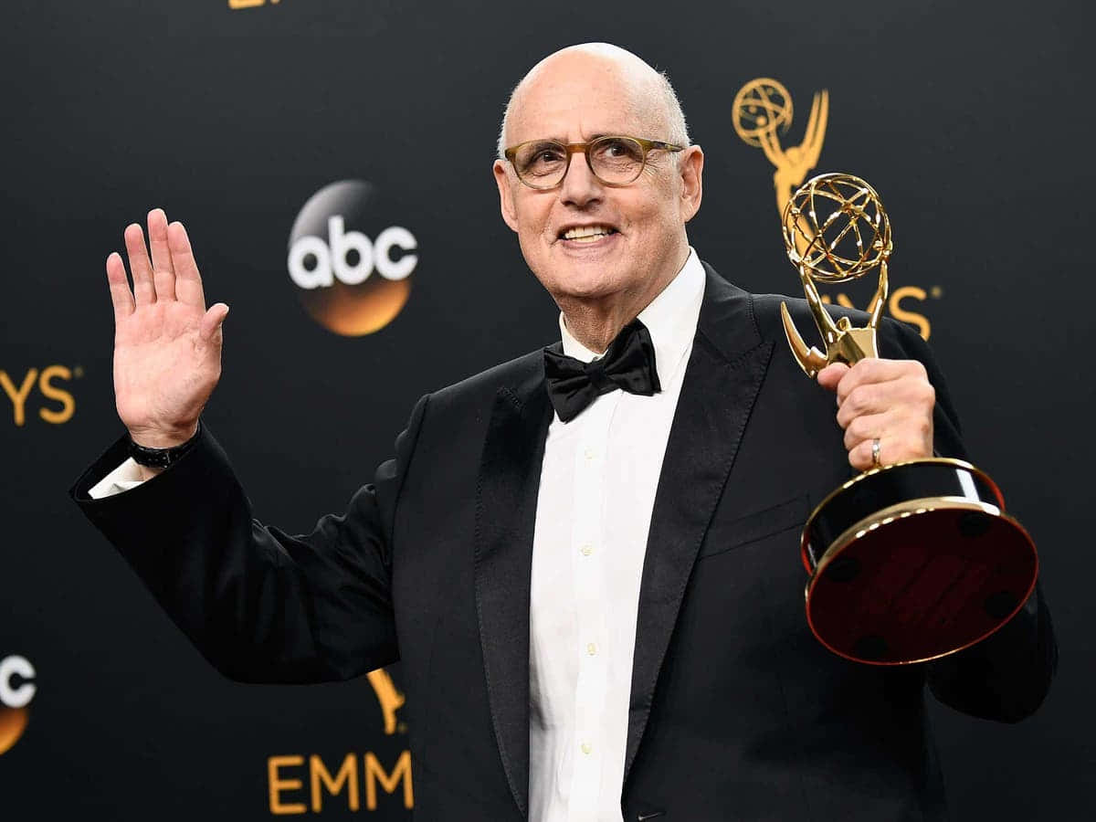 jeffrey tambor 桌布 壁紙 — 一個光頭男士穿著禮服，在黑色背景上有標誌，手持金色艾美獎並揮手。
