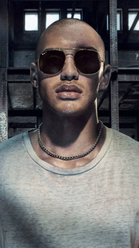 Sunglasses 桌布 壁紙 — 一個光頭男子戴著飛行員太陽眼鏡和鏈條項鍊，在昏暗的工業倉庫中凝視著。
