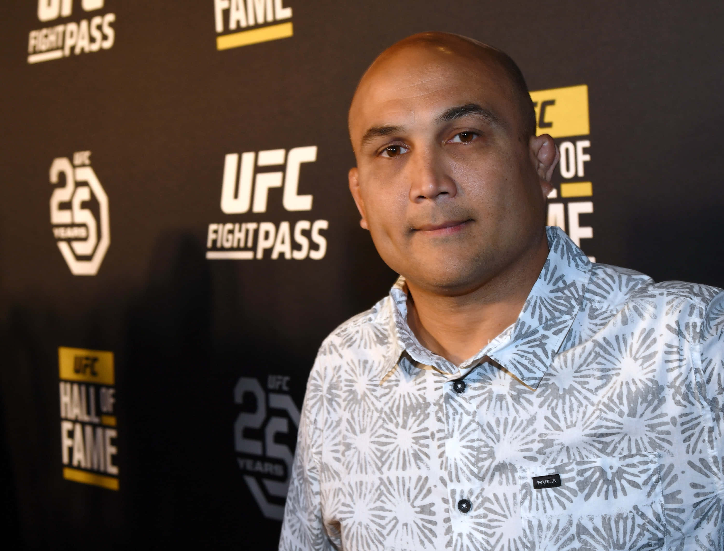 MMA 桌布 壁紙 — 一個光頭男子穿著圖案襯衫，自信地站在有品牌背景的UFC名人堂活動中。