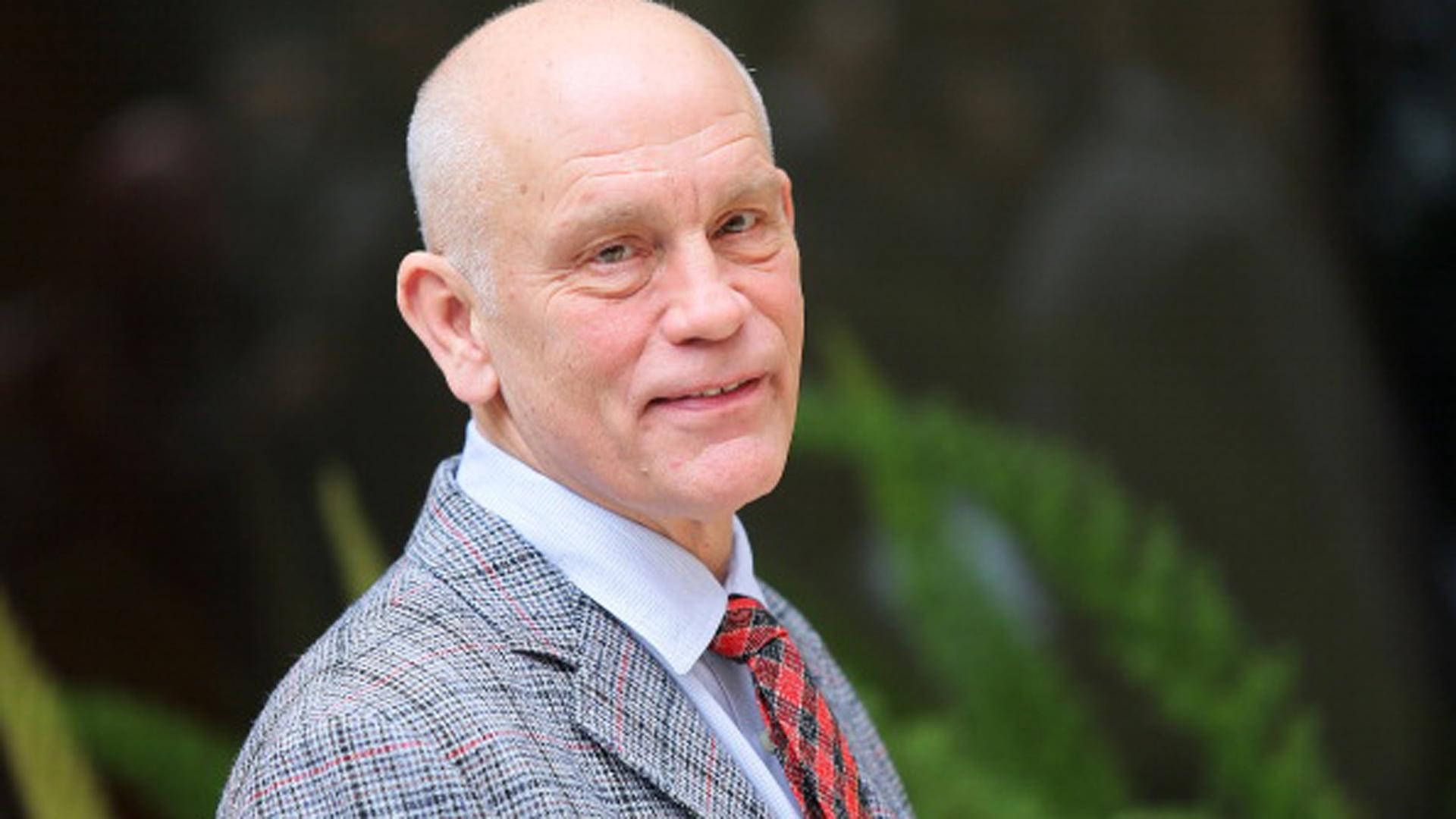 actor 桌布 壁紙 — 一個光頭男子穿著格紋西裝外套和紅色領帶，在模糊的綠色樹葉背景前溫柔地微笑。