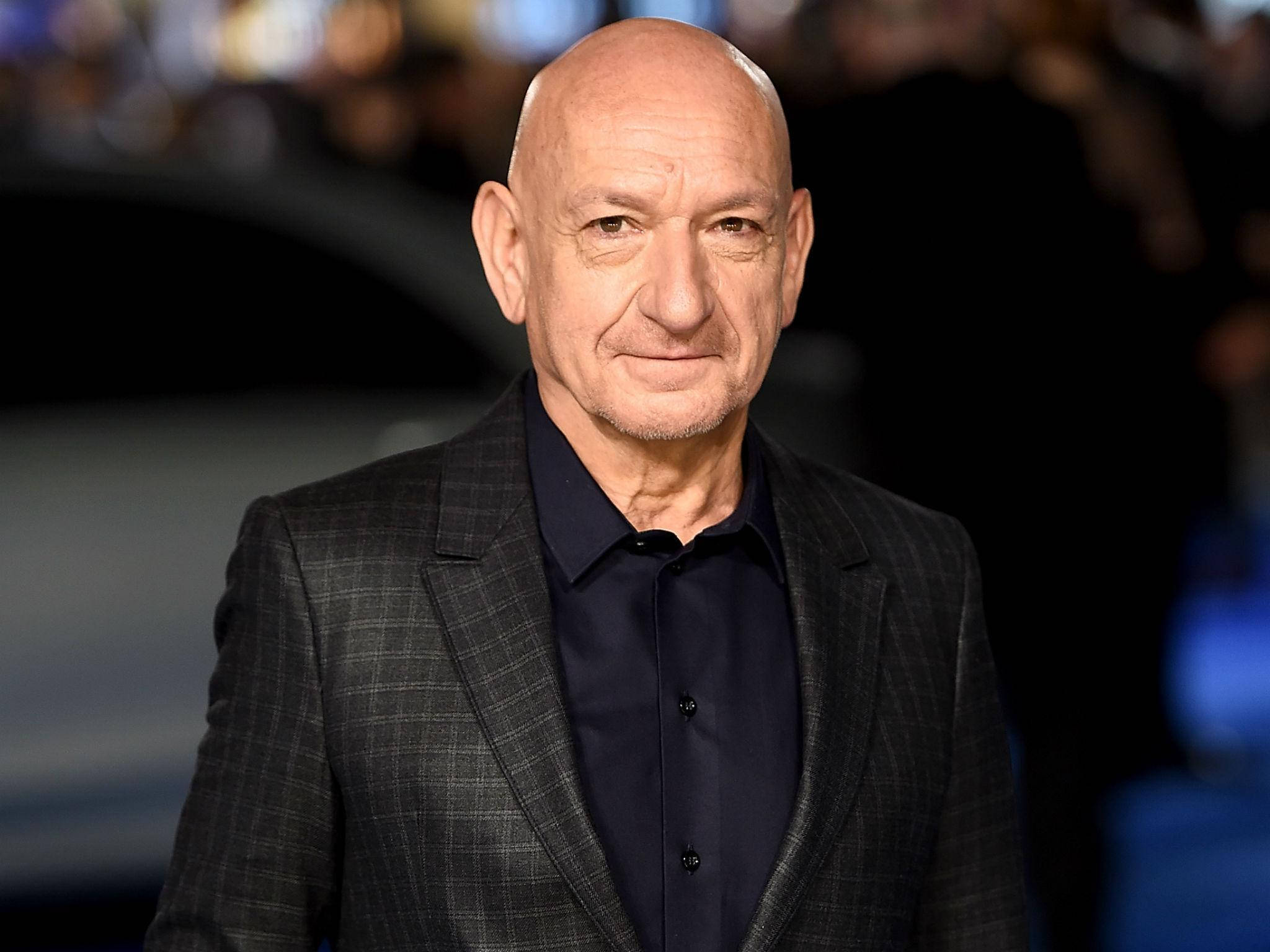 Celebrity 桌布 壁紙 — 一個光頭男子穿著深色格紋西裝和深藍色襯衫，在模糊的活動背景前對著鏡頭微笑。