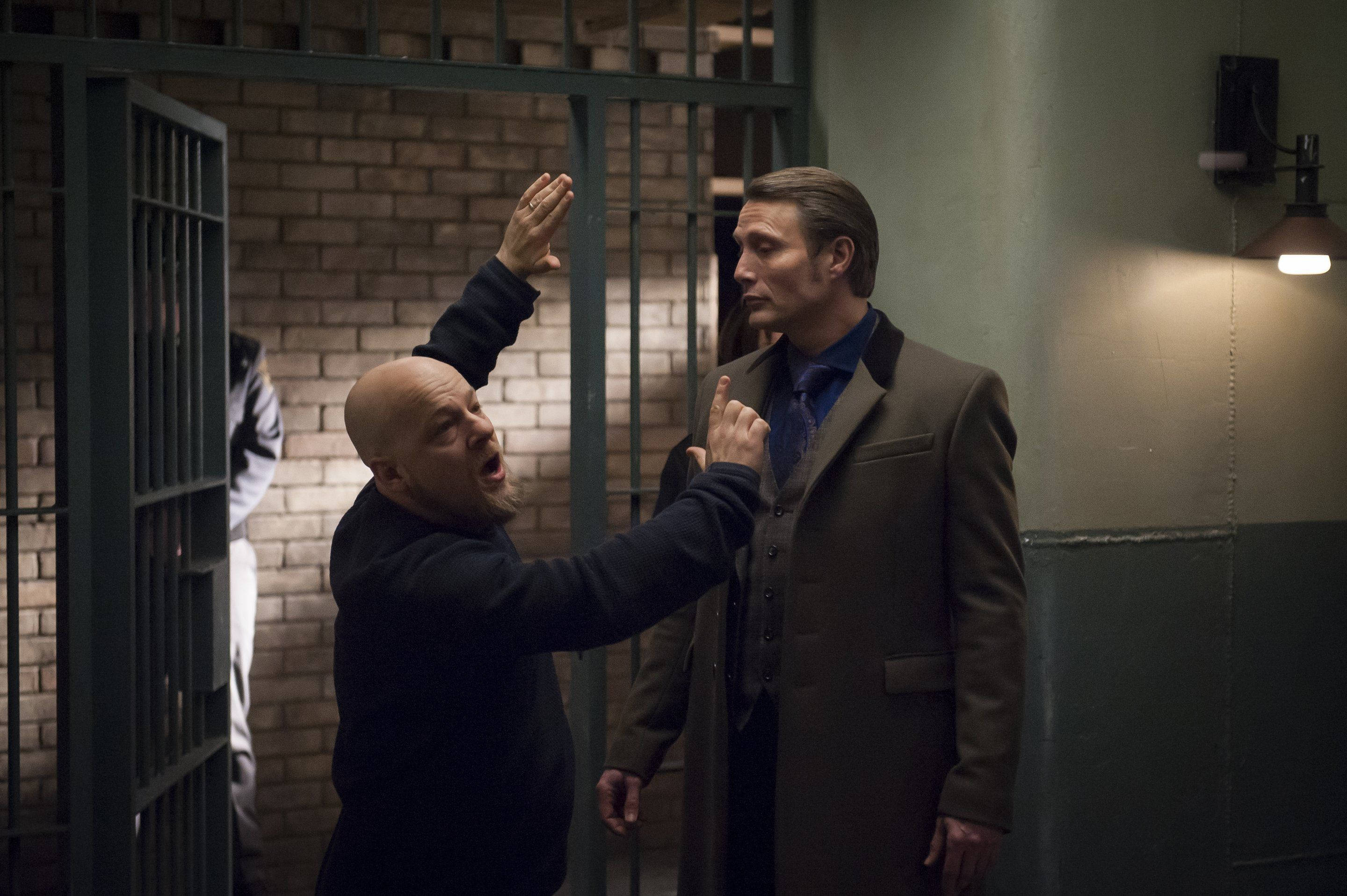 Hannibal 桌布 壁紙 — 一個光頭男子穿著深色襯衫，對著一個站在磚牆監獄牢房前的冷靜男子（身穿棕色外套）大喊大叫並激烈地做手勢。