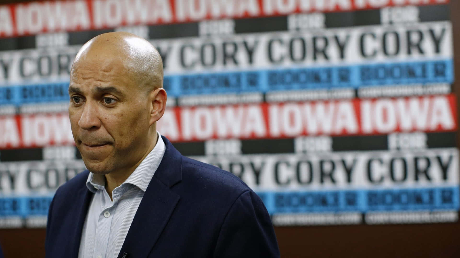 政治 桌布 壁紙 — 一個光頭男子穿著深色西裝，站在重複出現的「FOR CORY BIDEN」競選標語背景前。