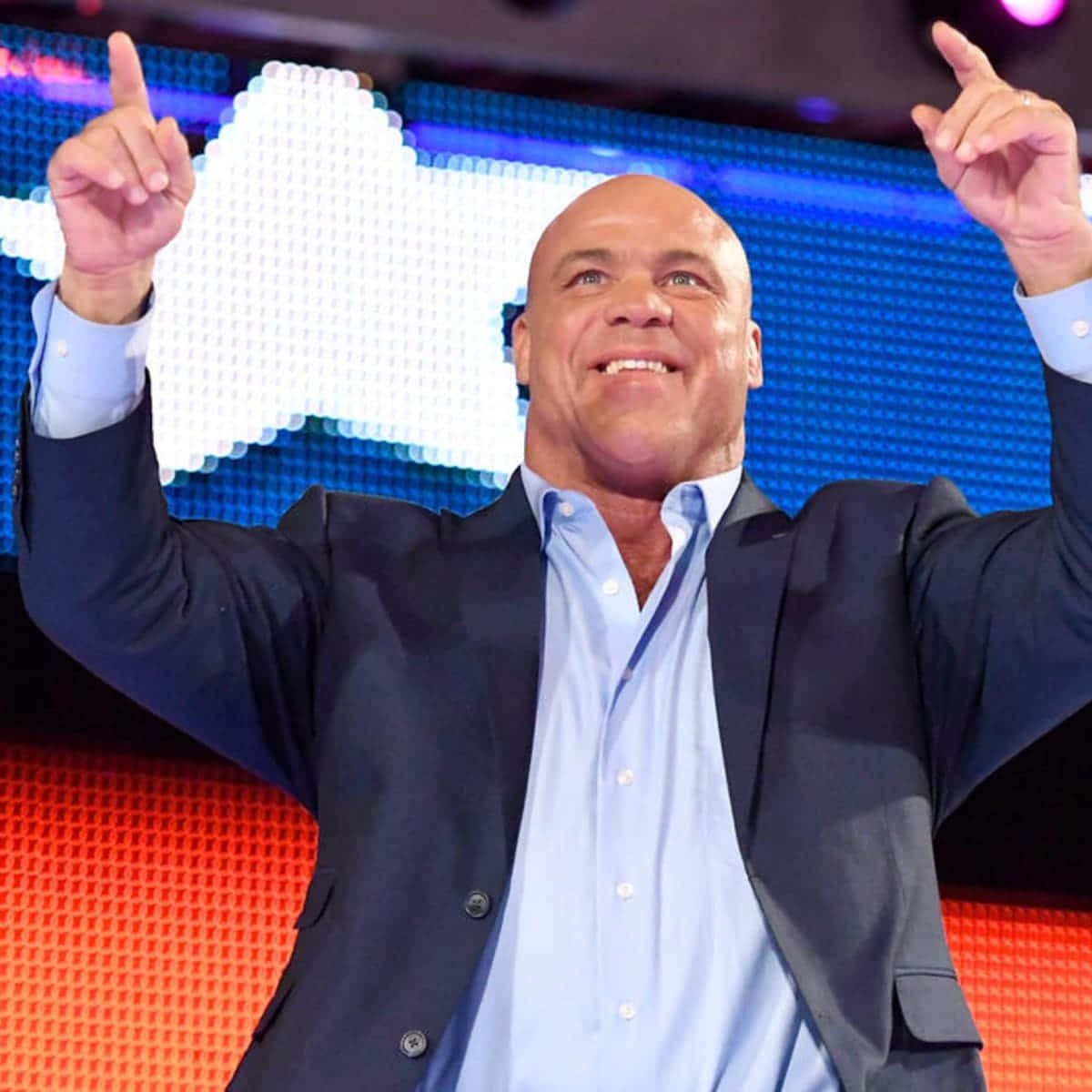 WWE 桌布 壁紙 — 一個光頭男子穿著深色西裝外套和淺藍色襯衫，在明亮的舞台上微笑，同時雙手指向上方，背景是像素化的紅色和藍色LED牆。