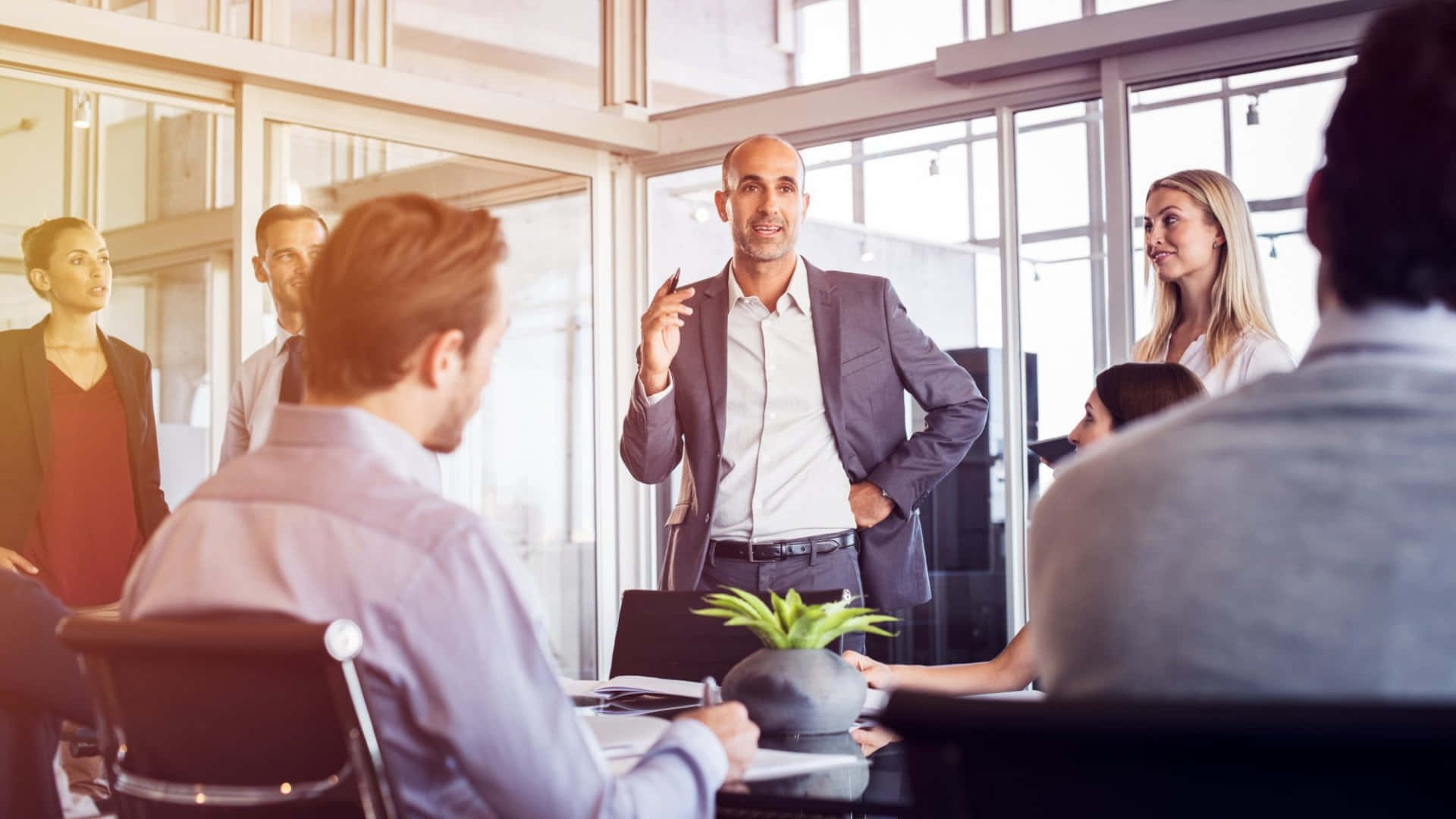 Leadership 桌布 壁紙 — 一個光頭男子穿著灰色西裝，在現代玻璃牆辦公室中向同事做簡報時做手勢。
