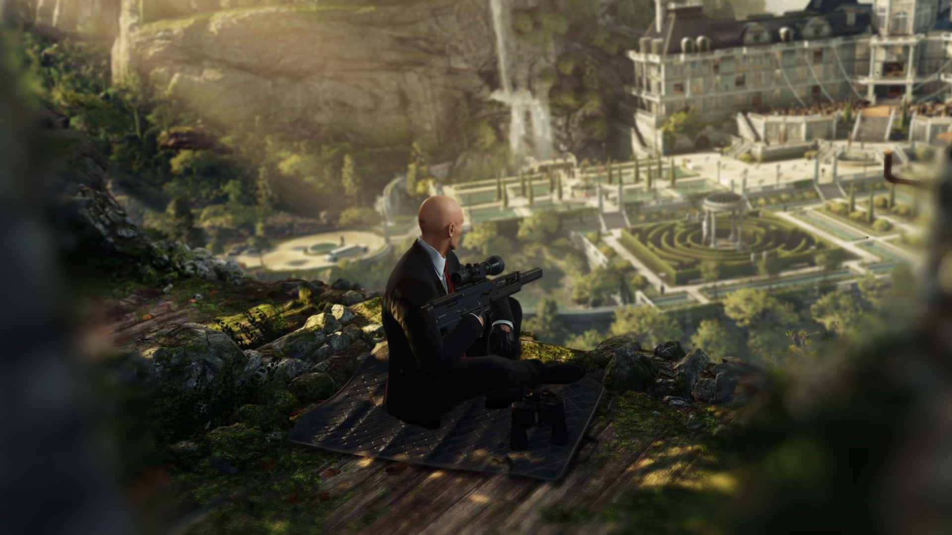 1920x1080 Hitman 2 桌布 壁紙 — 一個光頭男子穿著西裝坐在懸崖頂上，俯瞰著一個宏偉的莊園，有瀑布和修剪整齊的花園沐浴在金色的陽光下。