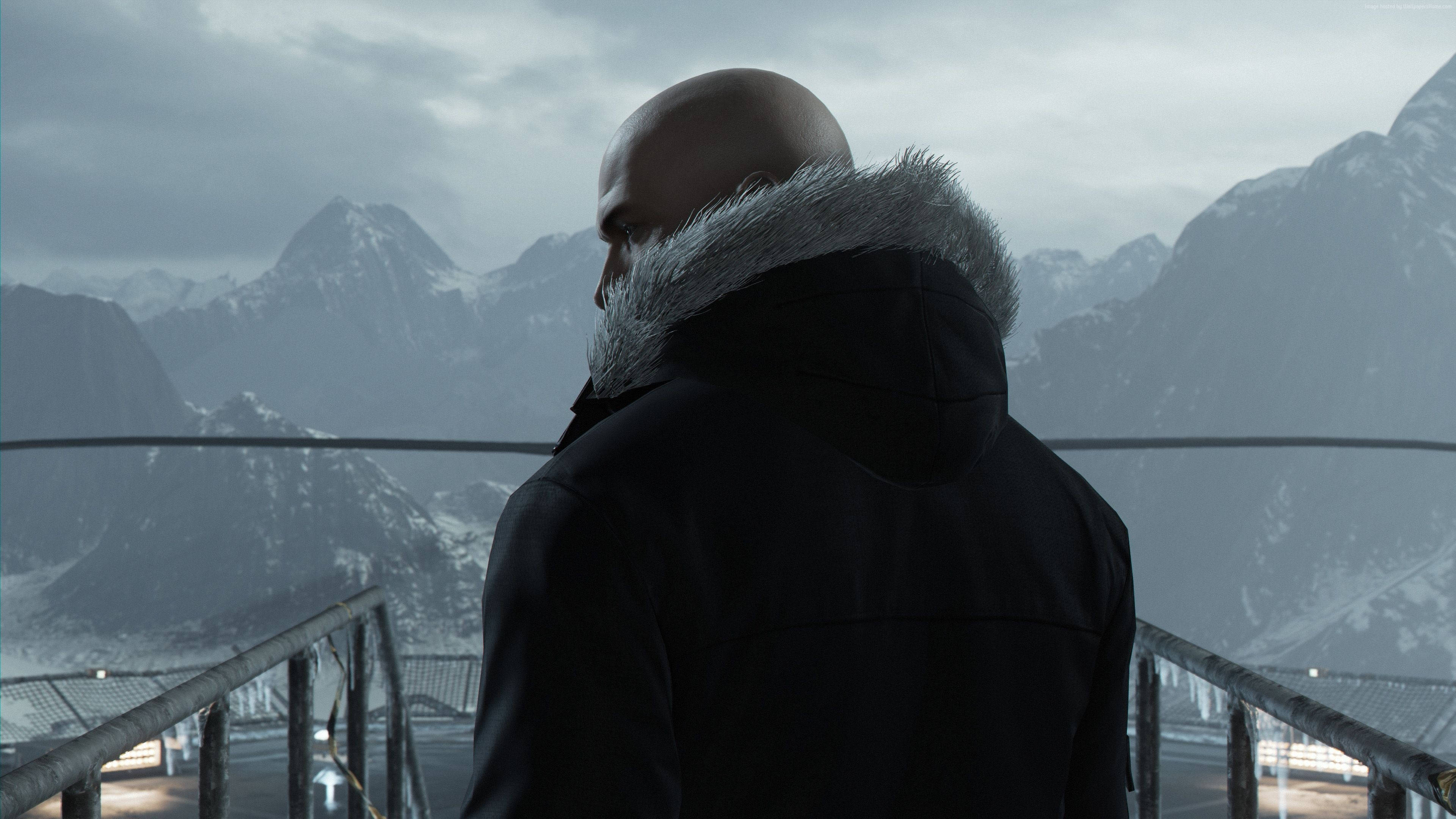 hitman absolution HD 桌布 壁紙 — 一個光頭男子穿著黑色外套，戴著毛皮襯裡的兜帽，站在金屬陽台上，俯瞰著多雲天空下的雪山。