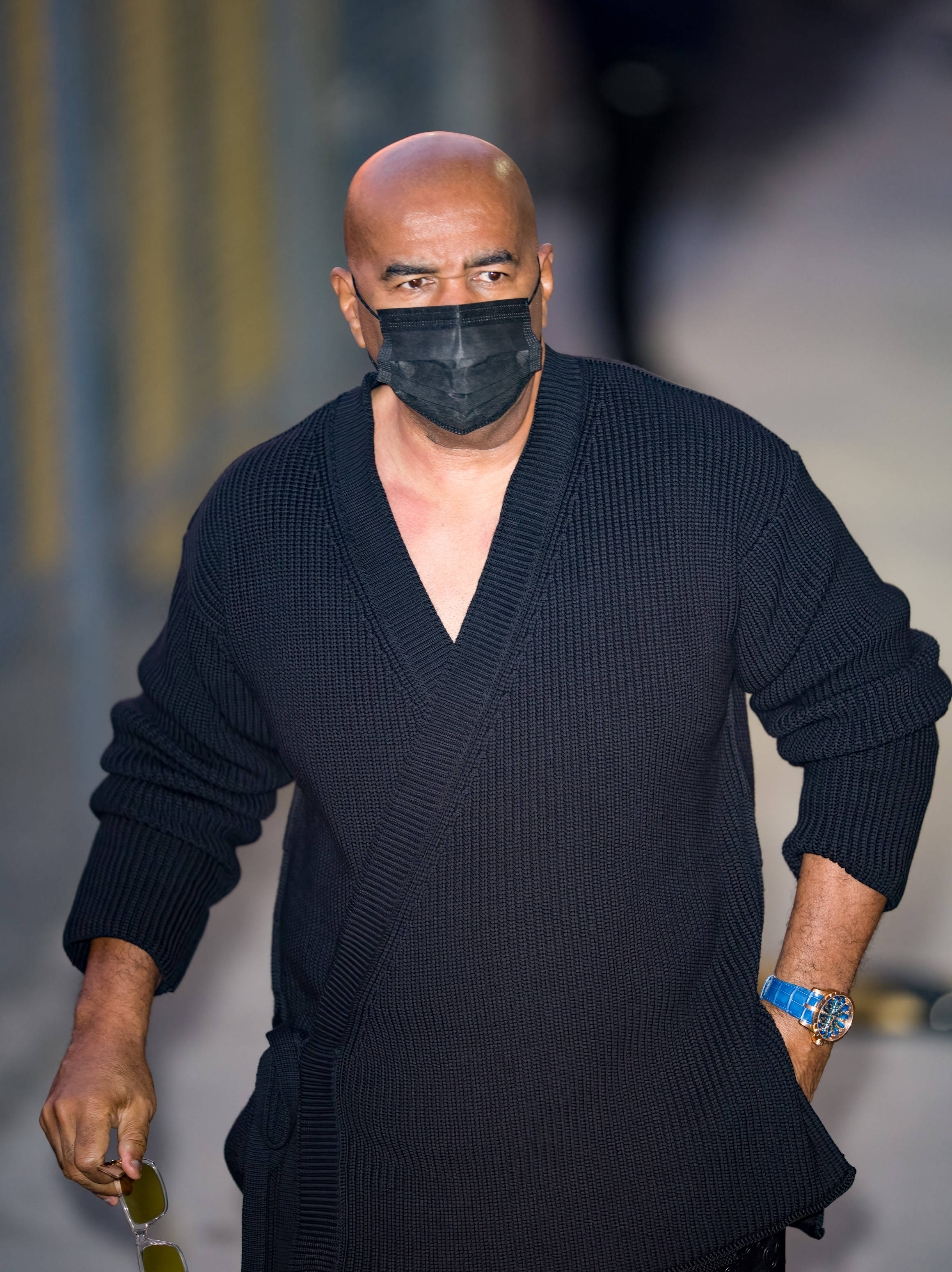 Celebrity 桌布 壁紙 — 一個光頭男子穿著黑色紋理開襟衫和口罩，自信地走在一條模糊的城市街道上，手持太陽眼鏡。