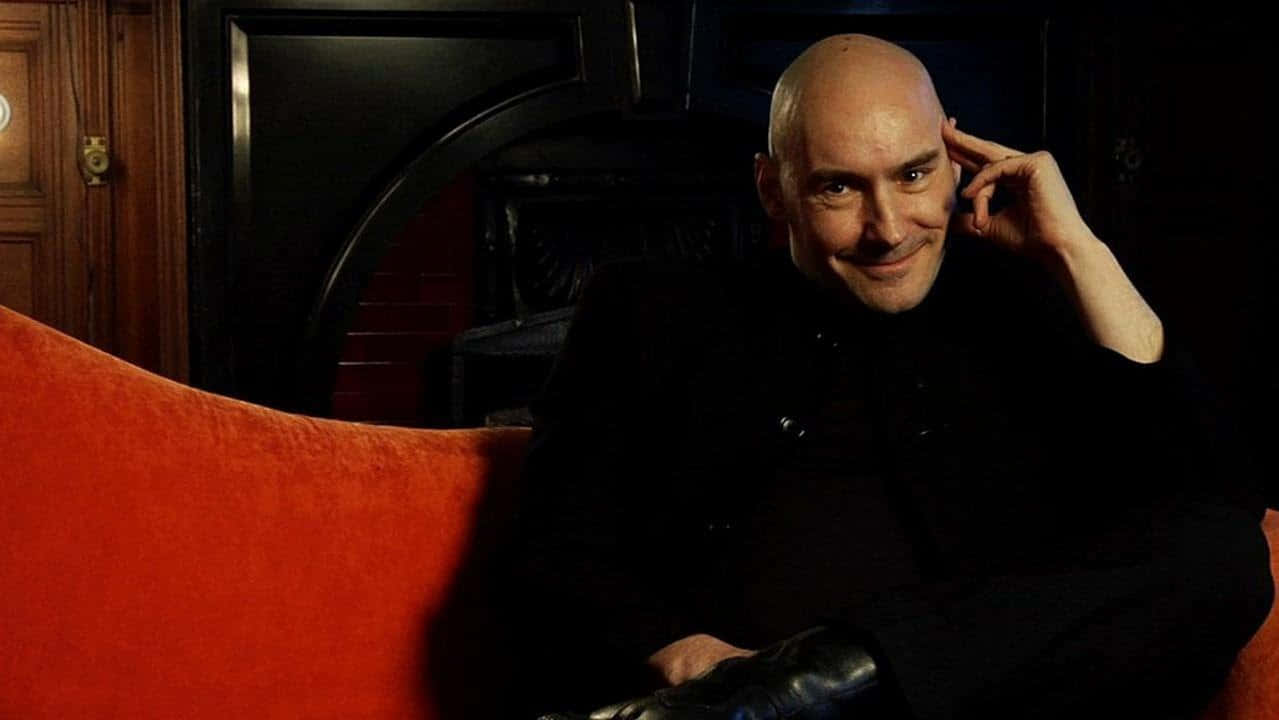 Grant Morrison 桌布 壁紙 — 一個光頭男子穿著黑色西裝，微笑著坐在一張柔軟的紅色沙發上，身後是昏暗的房間，有一扇木門和壁爐。