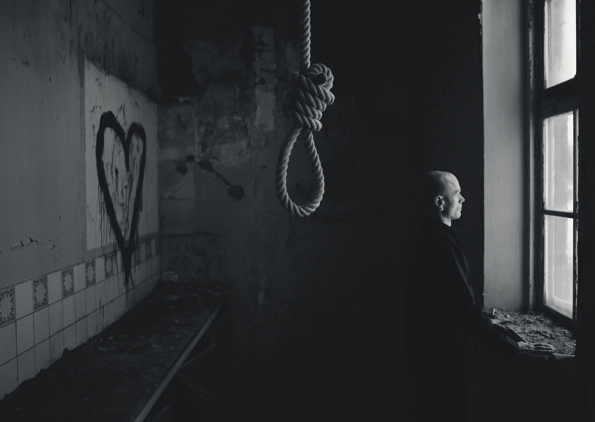 Life And Death 桌布 壁紙 — 一個光頭男子站在陰暗廢墟中窗邊，天花板上一個絞索在塗鴉心形附近陰森地懸掛著。