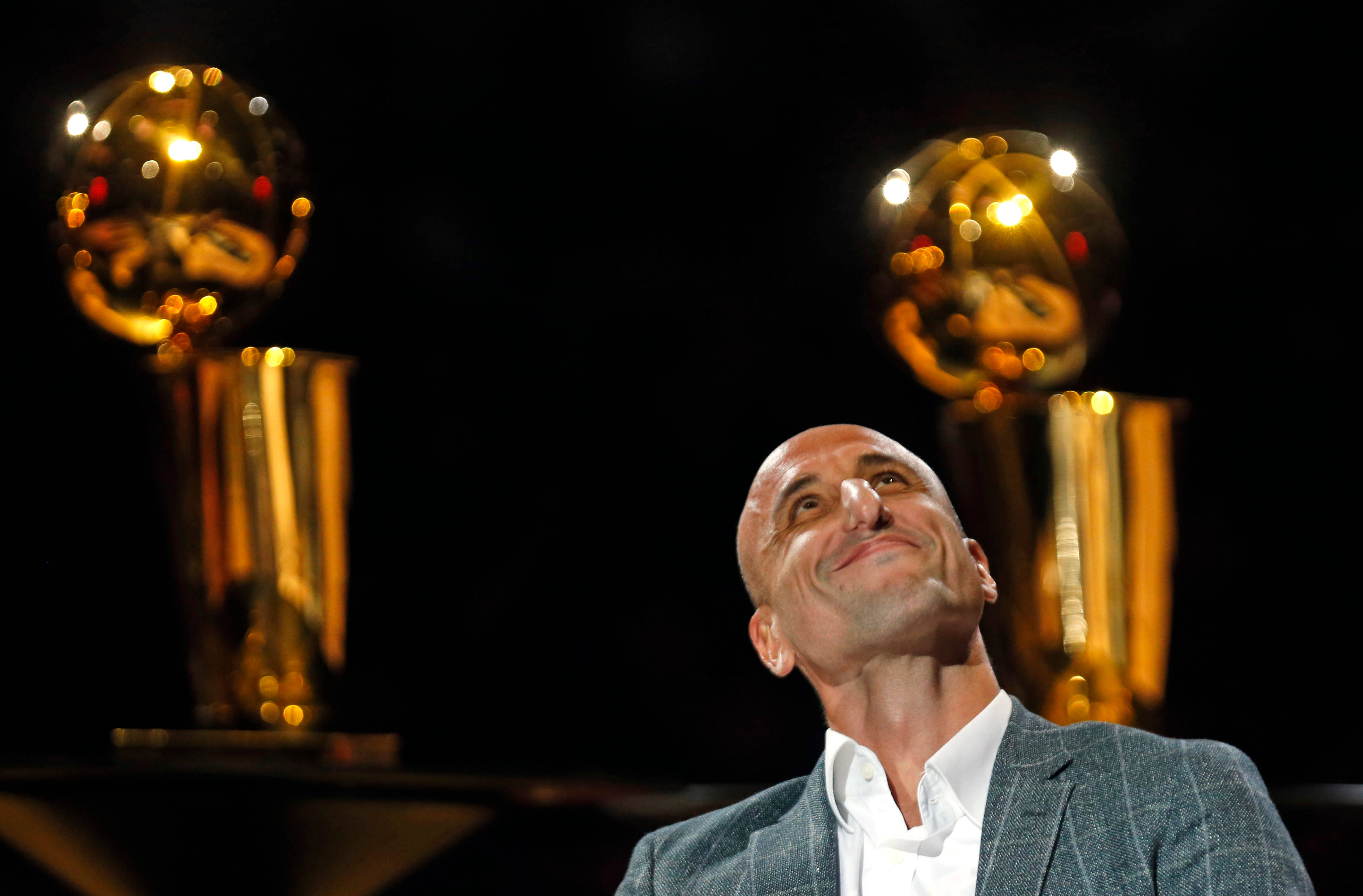Manu 桌布 壁紙 — 一個光頭男子西裝筆挺，向上微笑，背後有兩個金色的NBA冠軍獎盃在黑暗背景中發光。
