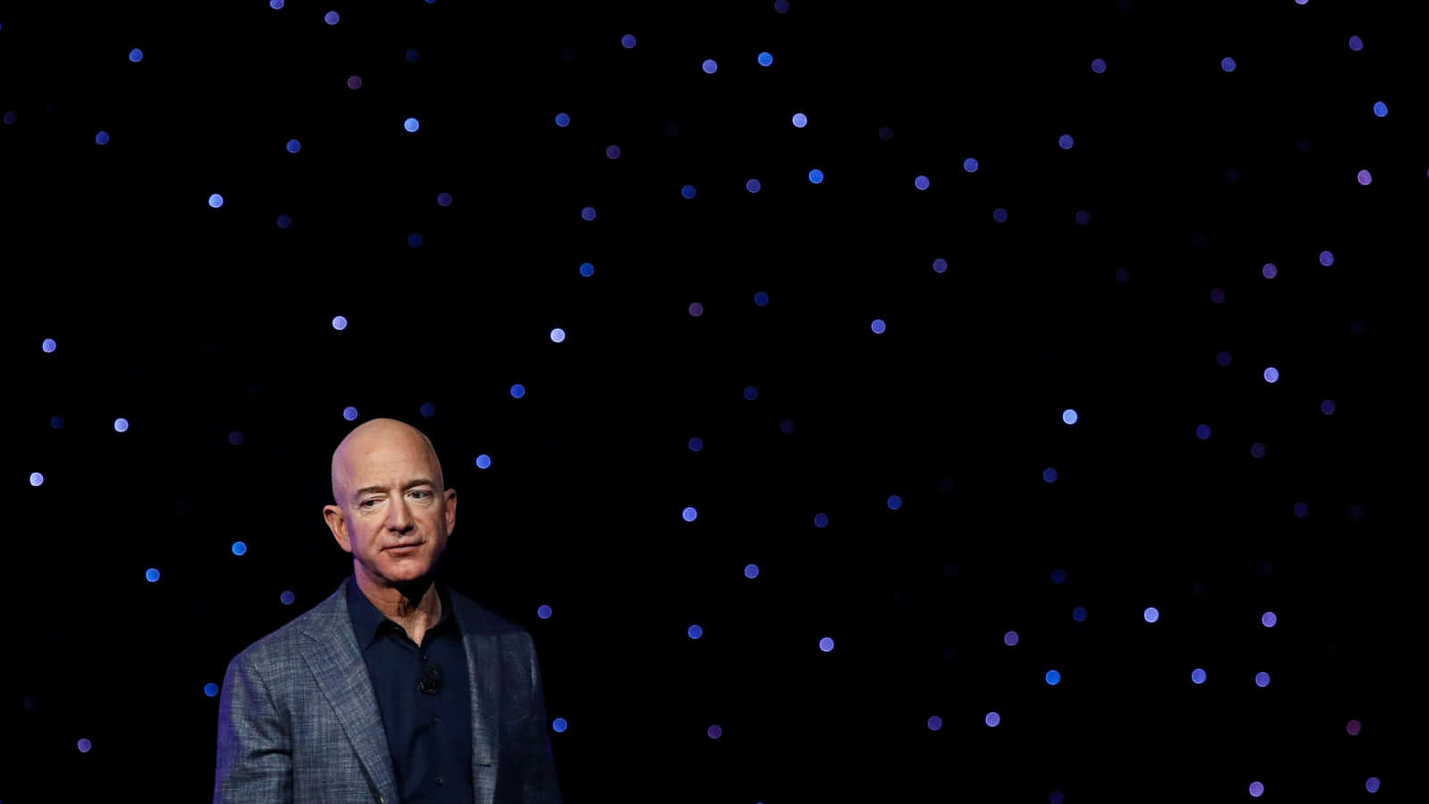 Jeff Bezos 桌布 壁紙 — 一個光頭男子身穿格紋西裝，自信地站在點綴著藍色光芒的黑色背景前。