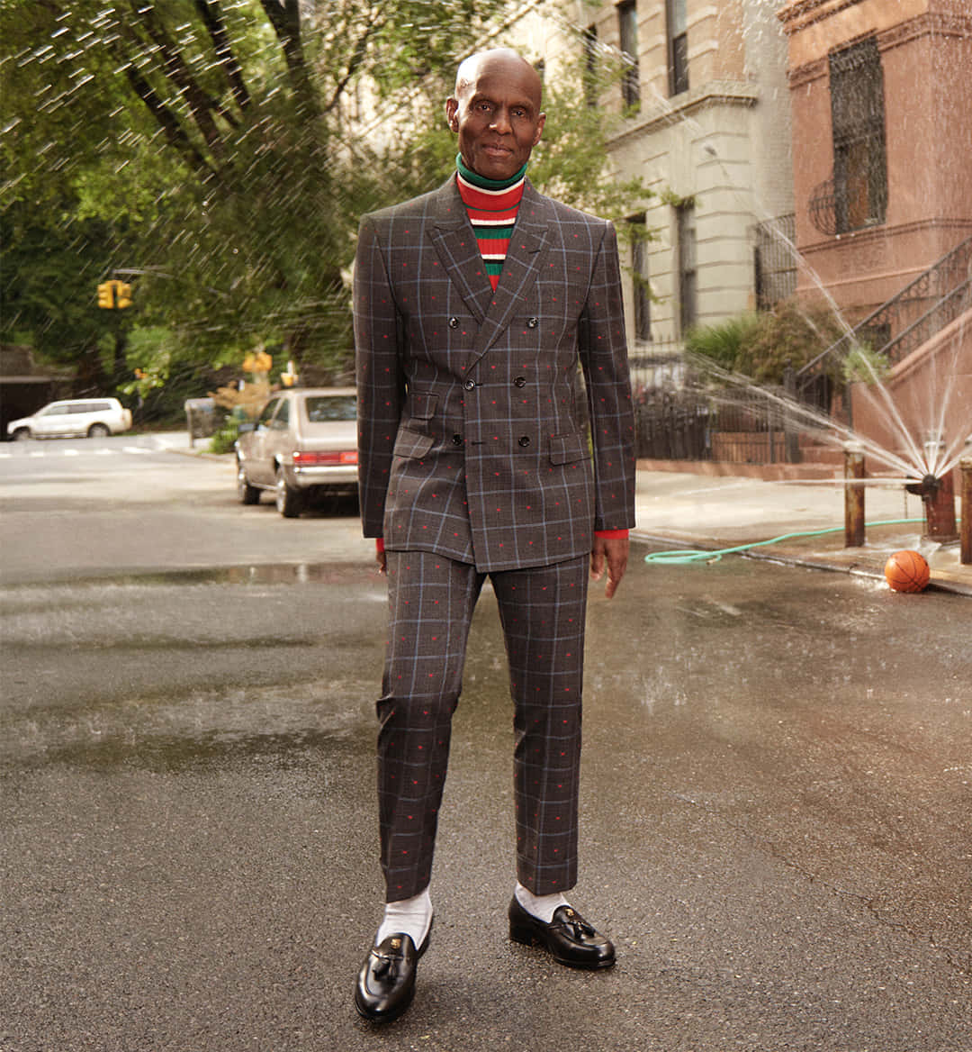 Man In Suit 桌布 壁紙 — 一個光頭男子身穿格紋西裝和條紋高領毛衣，自信地站在濕漉漉的城市街道上，附近的水管噴出水來。