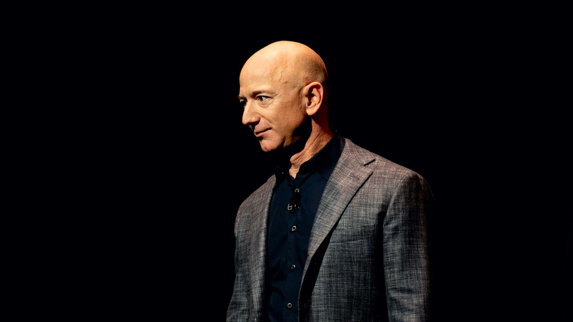 Jeff Bezos 桌布 壁紙 — 一個光頭男子身穿格紋西裝外套，在強烈黑色背景和戲劇性聚光燈下側面站立。