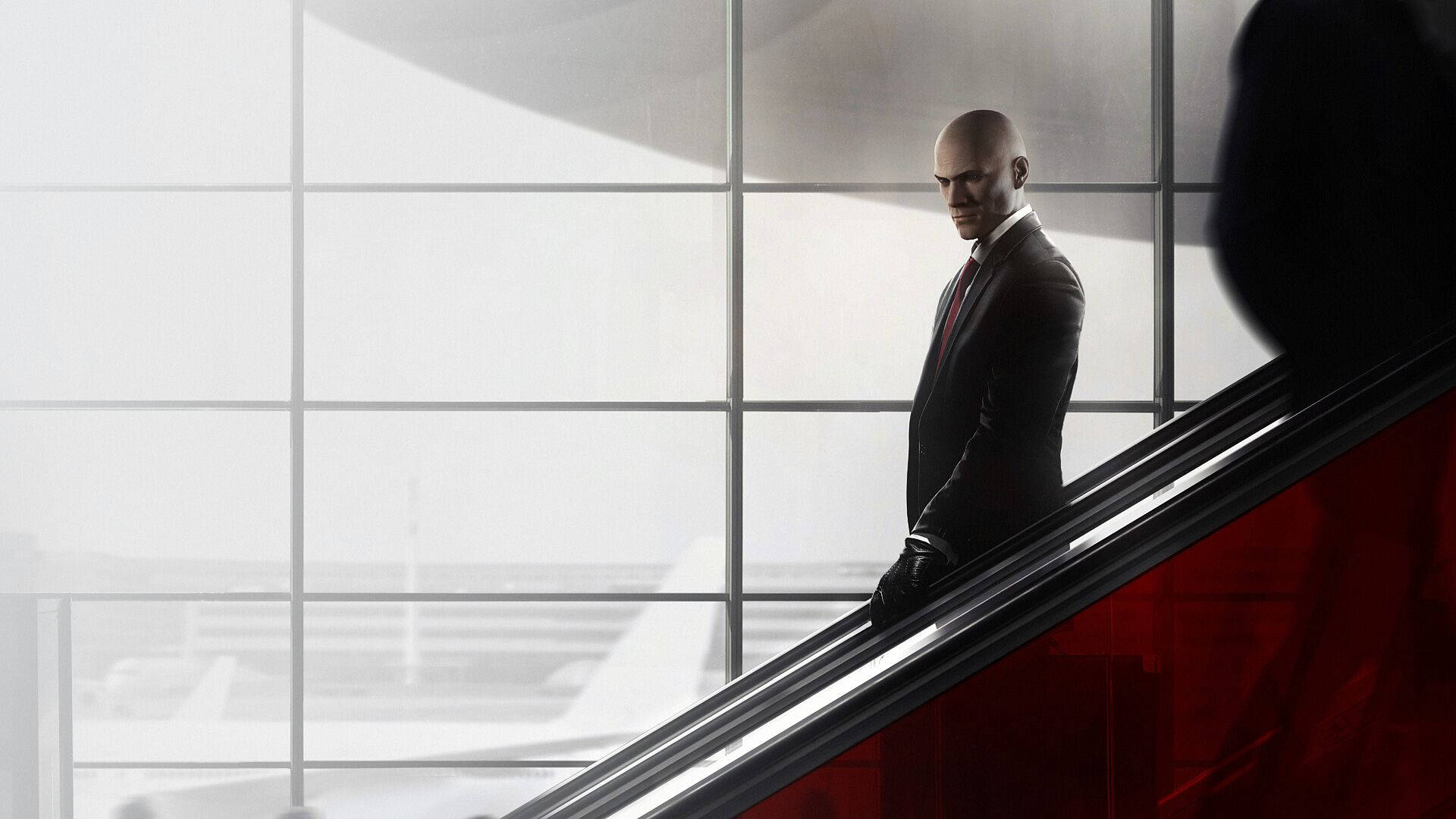 hitman absolution HD 桌布 壁紙 — 一個光頭男子身穿深色西裝，站在現代機場航廈的自動扶梯上，大窗戶和紅色點綴。