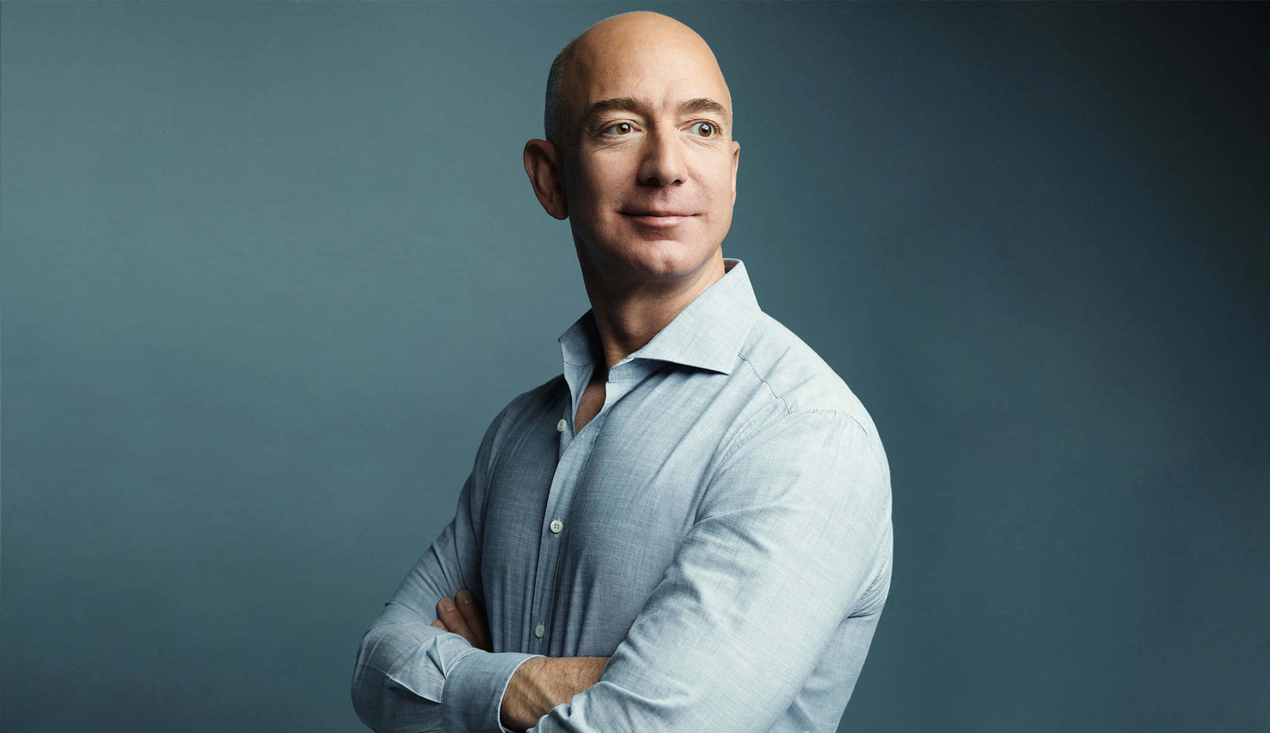 Jeff Bezos 桌布 壁紙 — 一個光頭男子身穿淺藍色襯衫，雙手交叉，自信地站在柔和的灰色背景前。