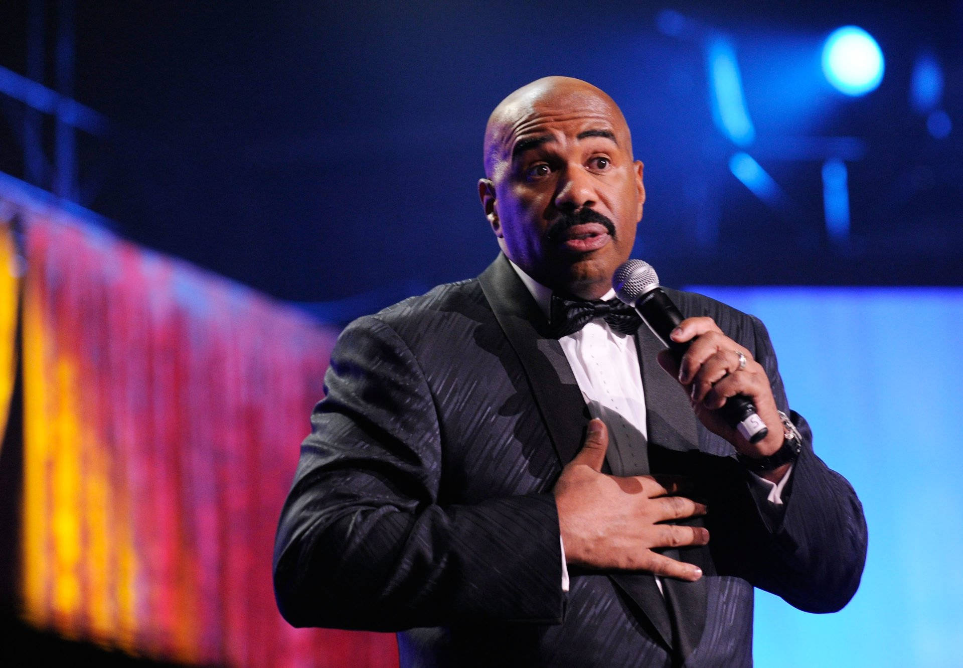 steve harvey 桌布 壁紙 — 一個光頭男子身穿黑色燕尾服配蝴蝶結，在舞台燈光下手持麥克風，另一隻手放在胸前做手勢。
