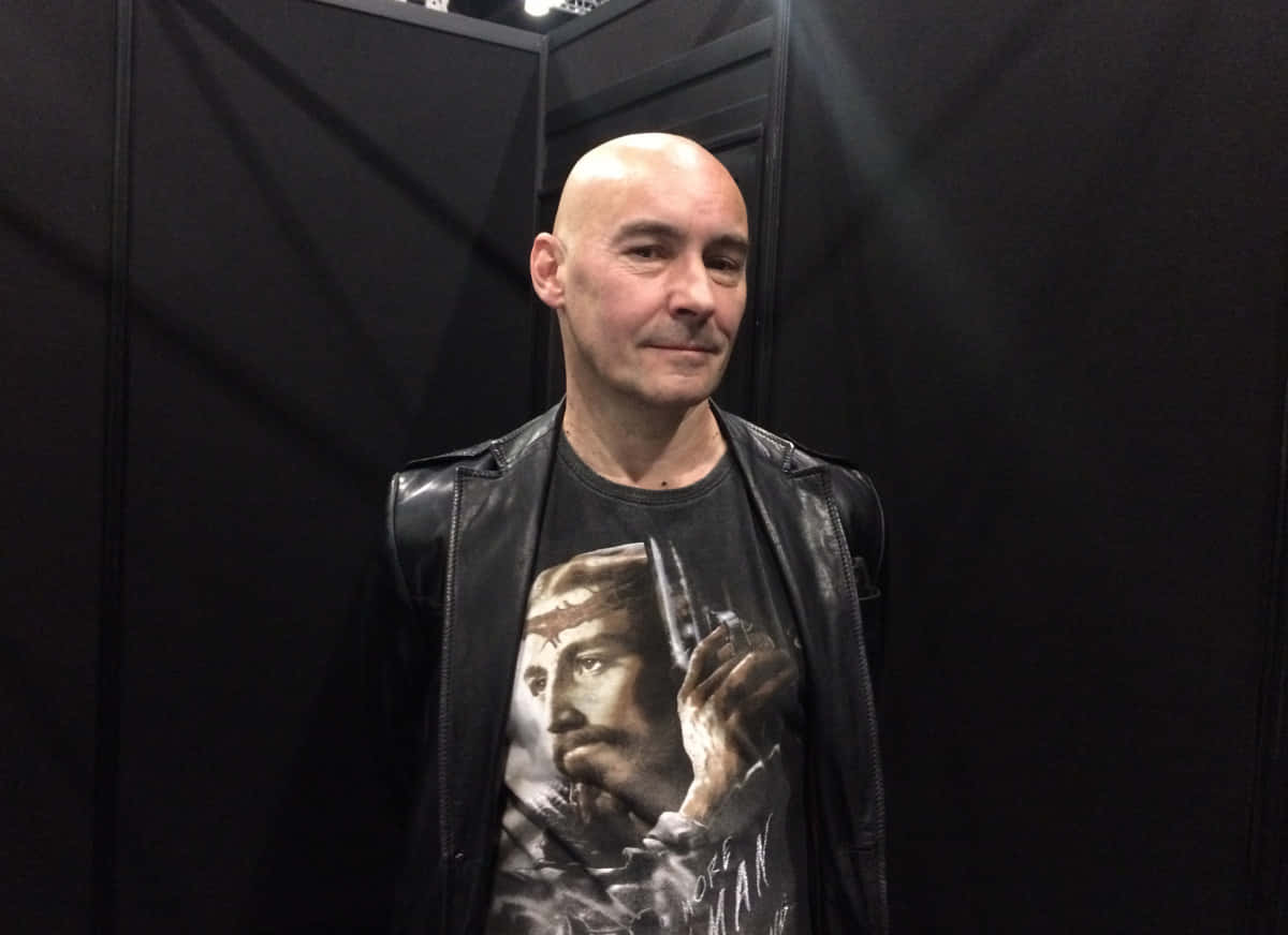 Grant Morrison 桌布 壁紙 — 一個光頭男子身穿黑色皮夾克和圖案T恤，站在柔和燈光的黑暗背景下。