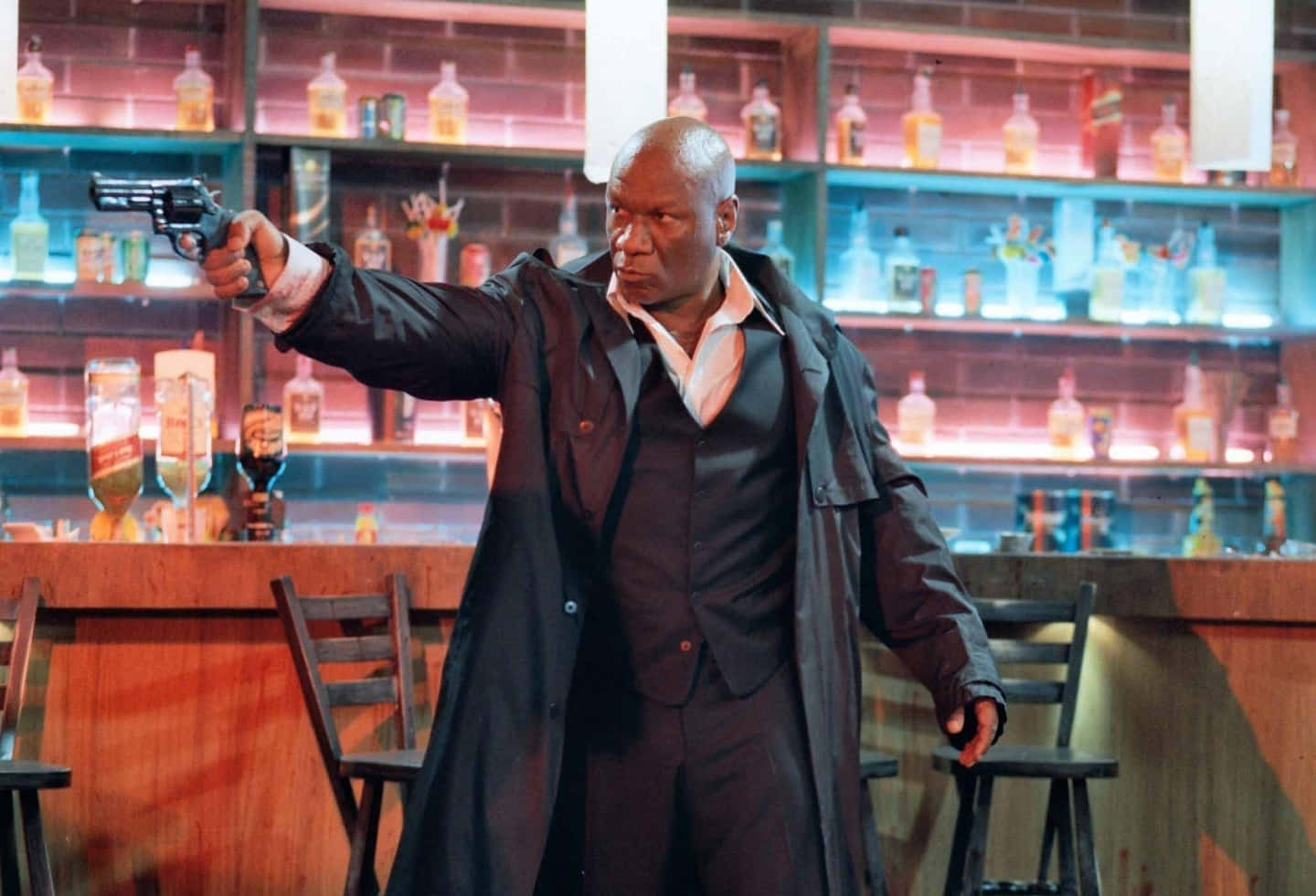 Ving Rhames 桌布 壁紙 — 一個光頭男子身穿黑色風衣，在燈光昏暗、身後有發光酒架的酒吧中瞄準左輪手槍。