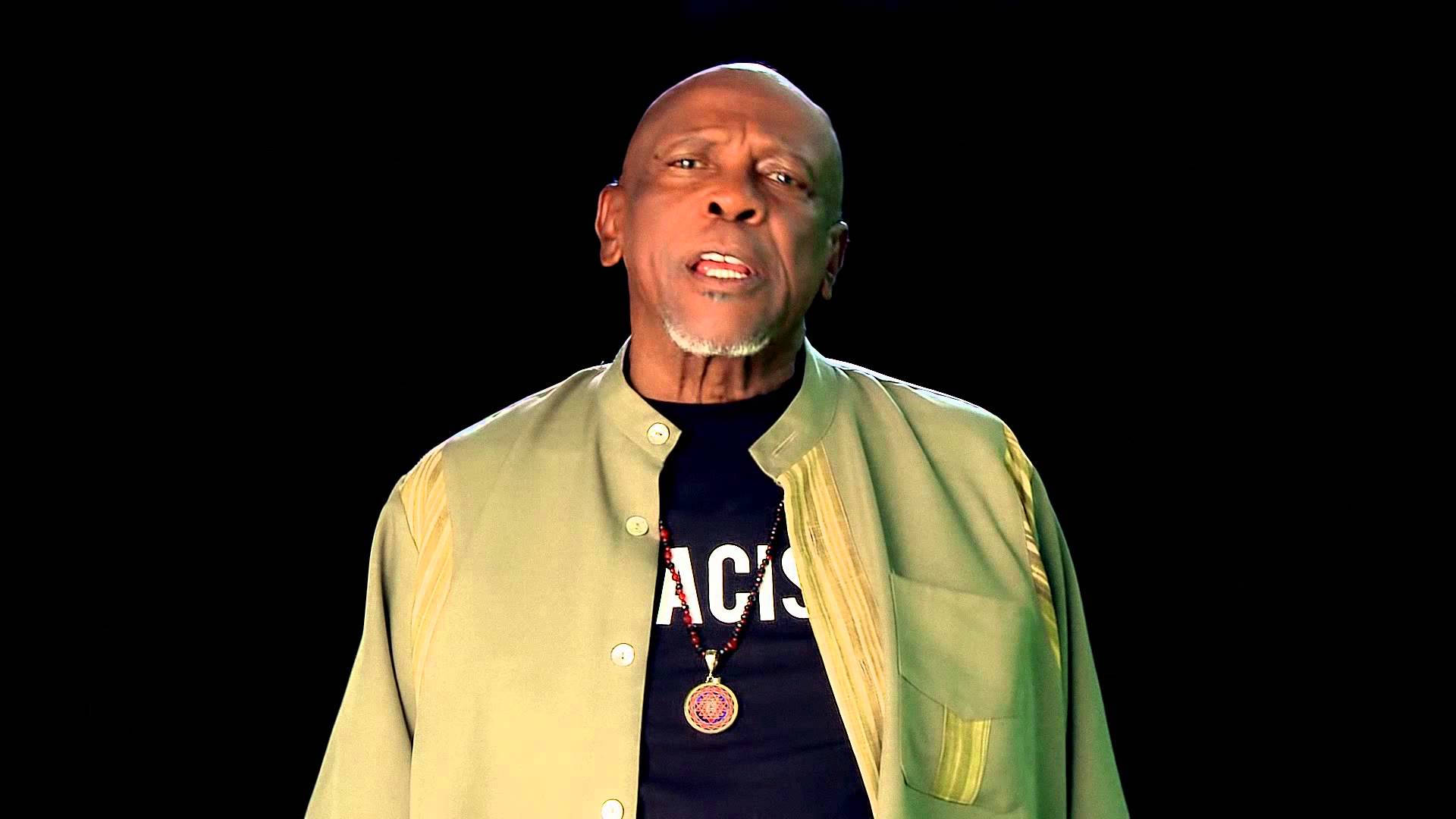 Celebrity 桌布 壁紙 — 一個光頭留山羊鬍的男人，在黑色背景前，穿著綠色外套，內搭黑色T恤，上面有白色文字和吊墜項鍊。