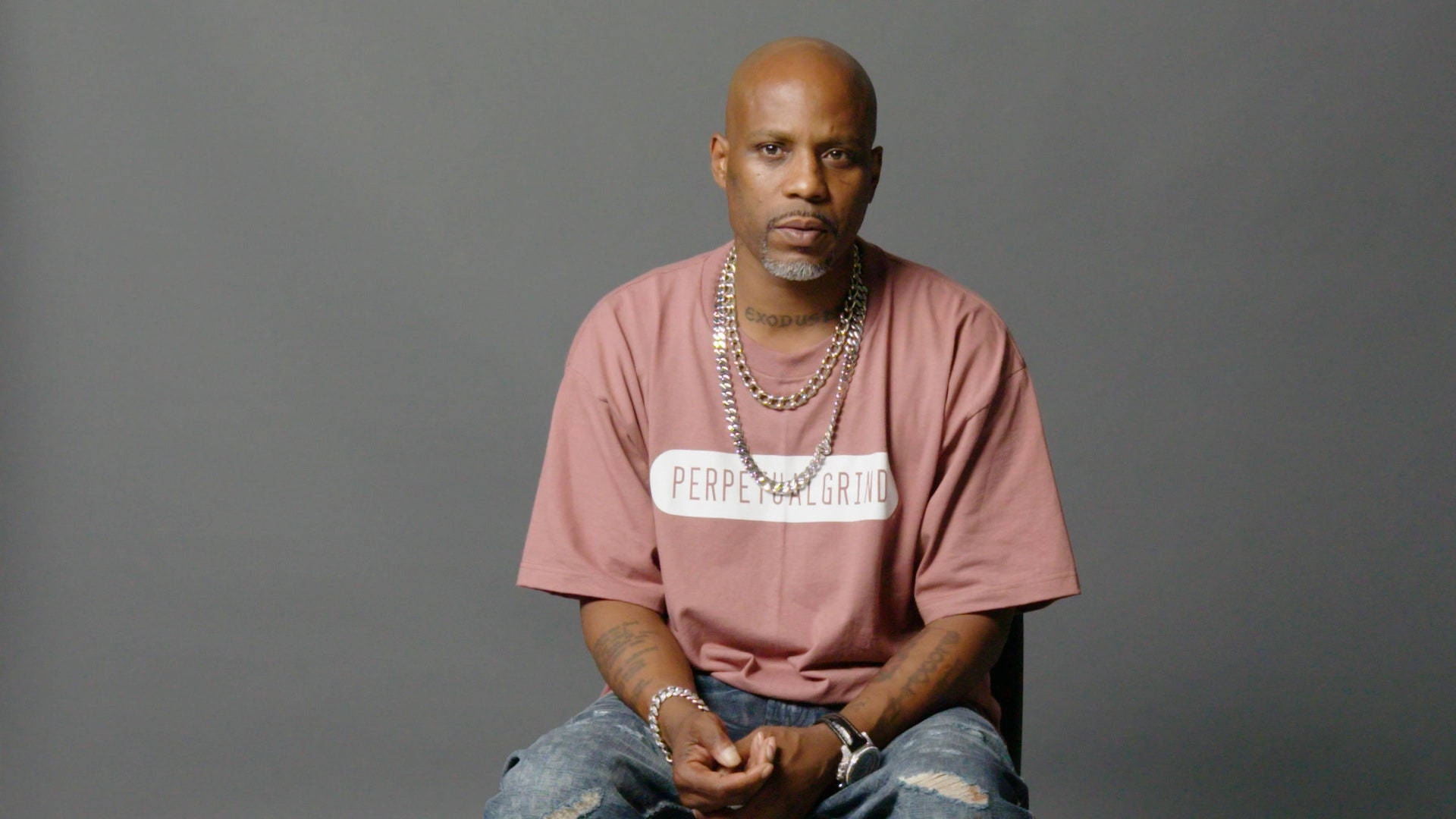 Dmx 桌布 壁紙 — 一個光頭留山羊鬍的男人平靜地坐在椅子上，穿著粉紅色T恤和層疊鏈條，背景是純灰色。