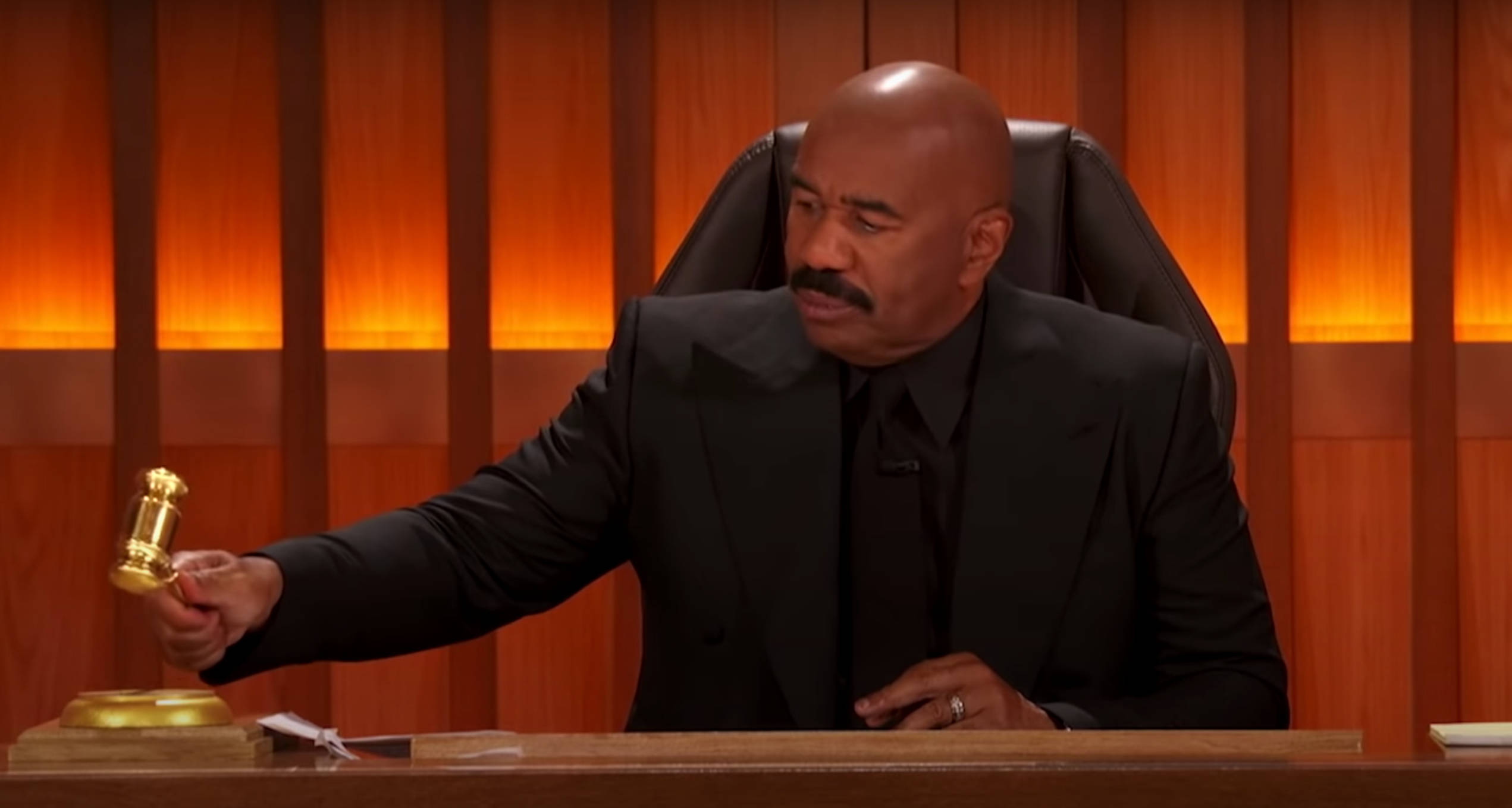 steve harvey 桌布 壁紙 — 一個光頭留鬍鬚的男人穿著黑色西裝，在溫暖燈光照射的木鑲板背景下，於木桌上握著金色木槌。